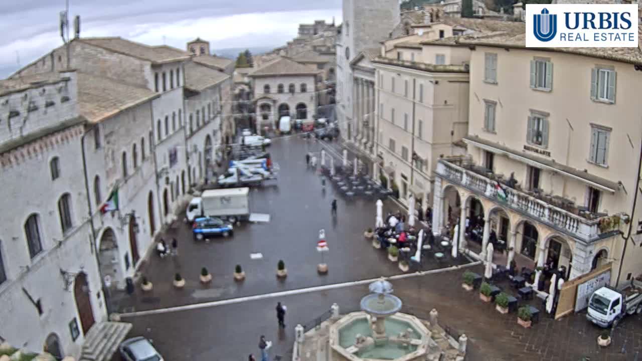 A historic European town square featuring a central fountain, surrounding buildings with outdoor cafes, and various people and vehicles on wet pavement beneath an overcast sky.