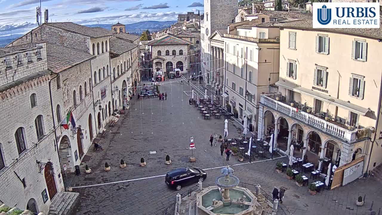 A historic European town square featuring a central fountain, surrounding buildings with outdoor cafes, and various people and vehicles on wet pavement beneath an overcast sky.