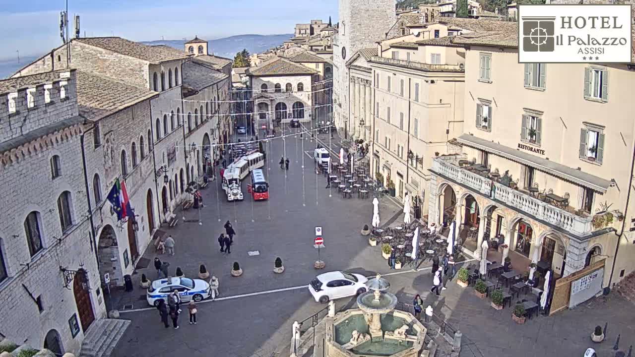 A vibrant European town square is bustling with pedestrians, cars, and outdoor dining under a clear, sunny sky, featuring historic stone buildings and a decorative fountain.