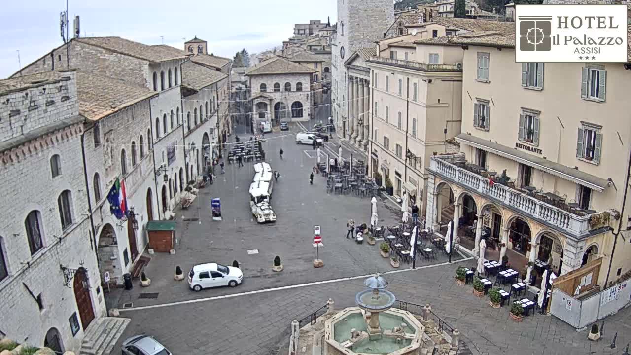 An overcast day illuminates a lively historic European town square featuring a central fountain, numerous outdoor restaurant patios with patrons, pedestrians, and various vehicles moving through or parked in the area.