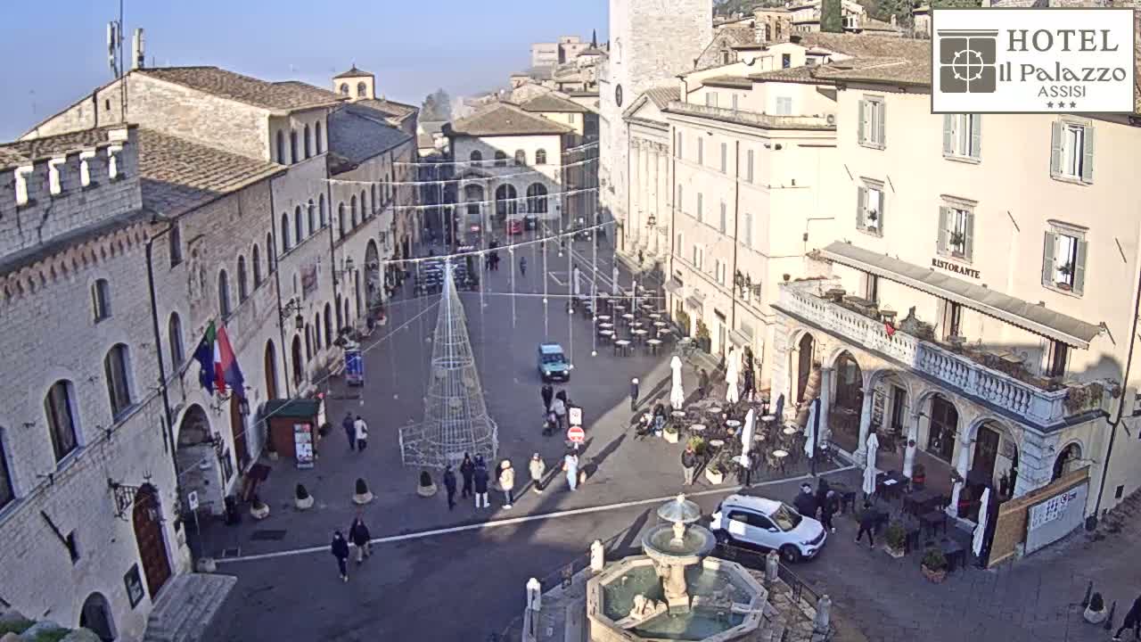 An overcast day illuminates a lively historic European town square featuring a central fountain, numerous outdoor restaurant patios with patrons, pedestrians, and various vehicles moving through or parked in the area.