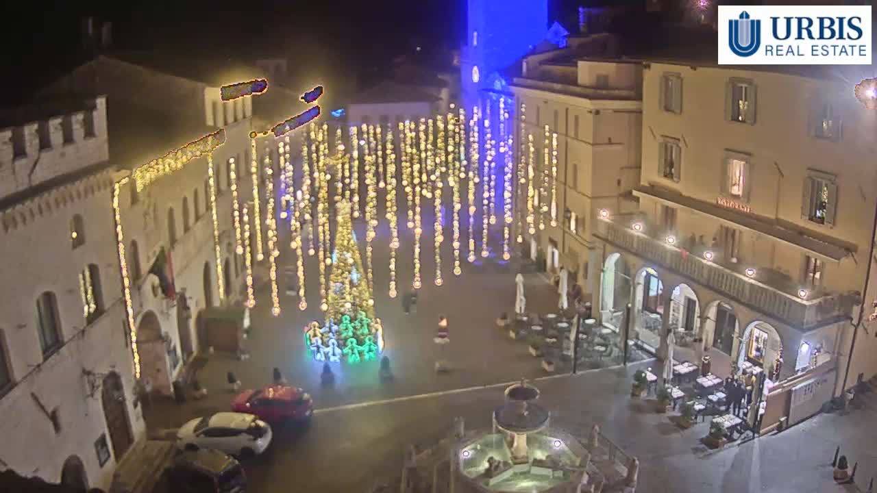 An overcast day illuminates a lively historic European town square featuring a central fountain, numerous outdoor restaurant patios with patrons, pedestrians, and various vehicles moving through or parked in the area.