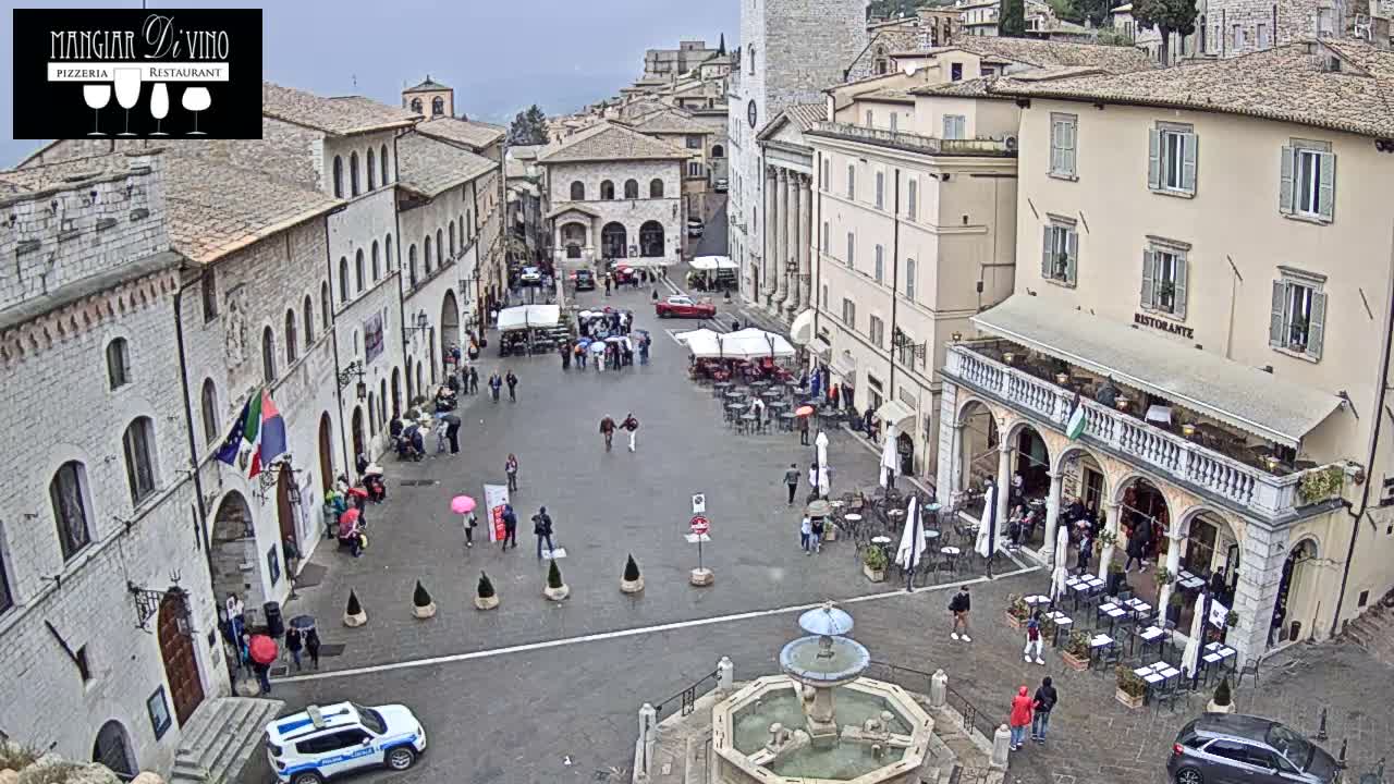 An overcast day illuminates a lively historic European town square featuring a central fountain, numerous outdoor restaurant patios with patrons, pedestrians, and various vehicles moving through or parked in the area.