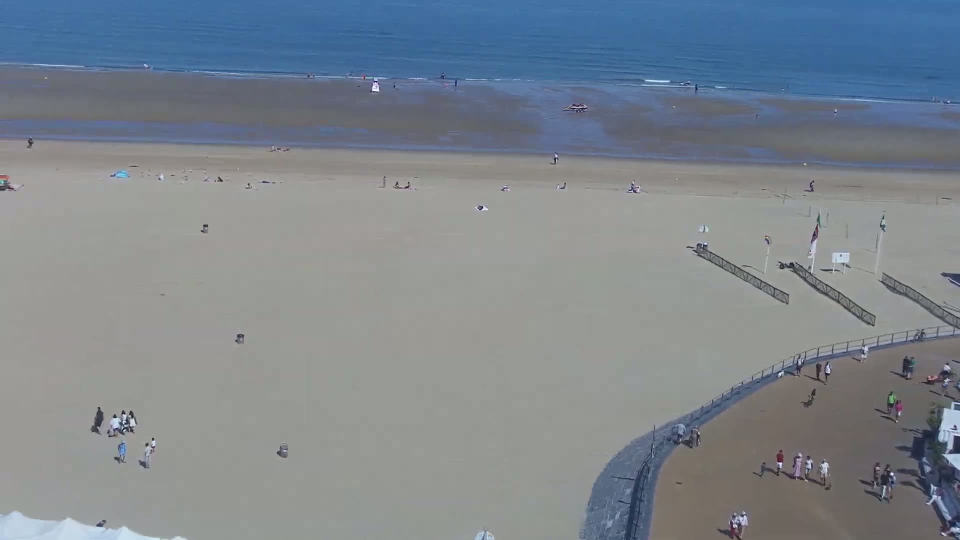 An aerial view shows a sandy beach with scattered people sunbathing and playing, adjacent to a paved area with more people, all under a sunny sky and calm ocean.