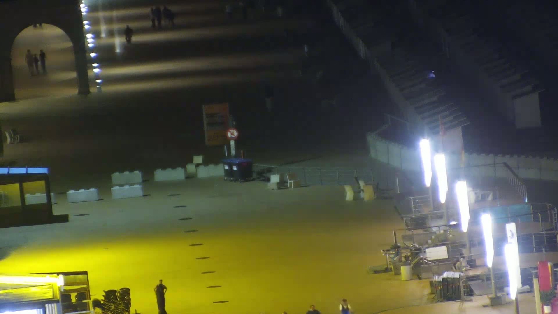 An illuminated nighttime scene shows an outdoor area with several people sparsely scattered, various structures, and barriers.