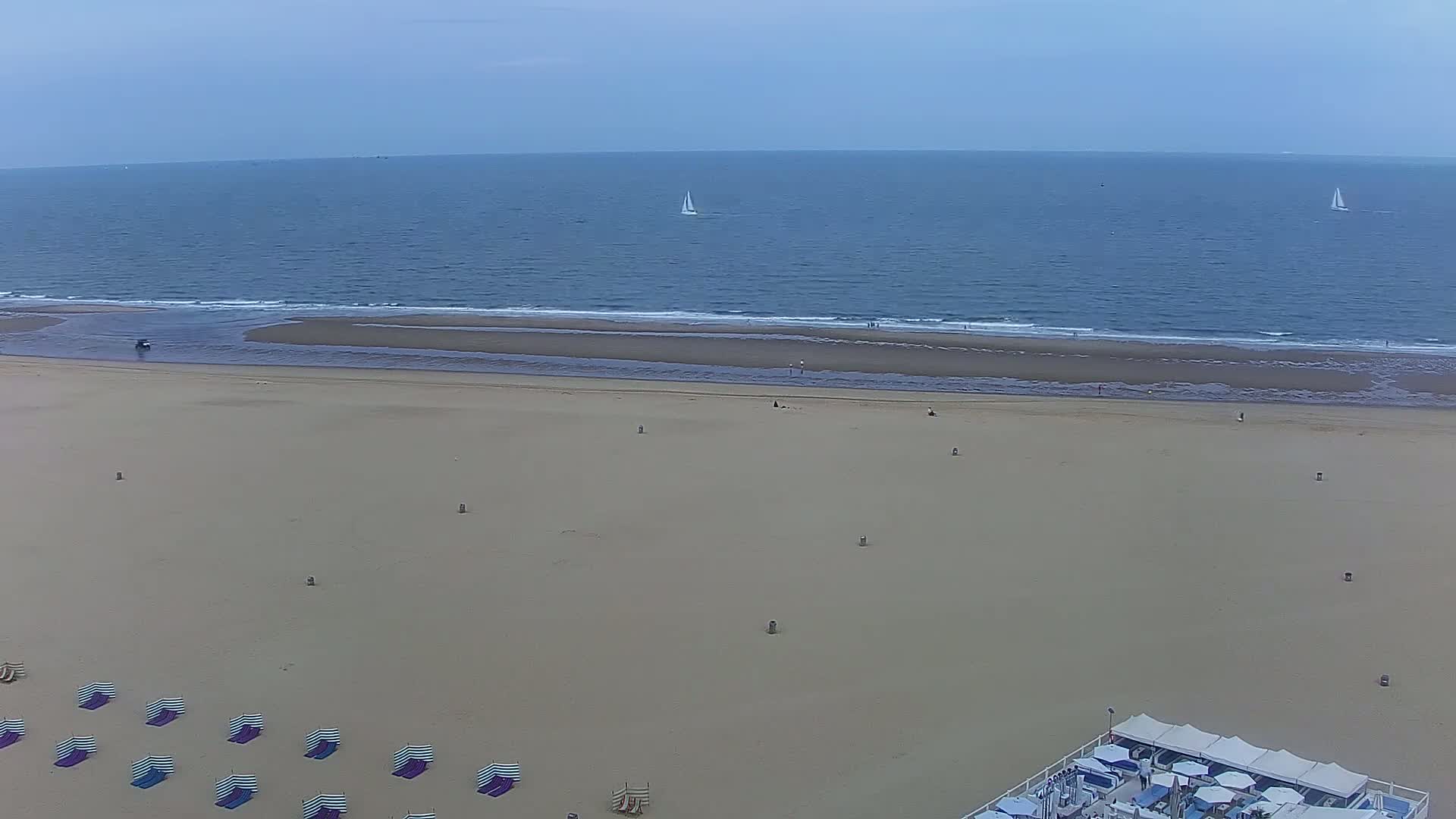 An aerial view shows a wide, sandy beach with a few people and striped beach umbrellas, next to a calm ocean with two sailboats visible in the distance under a cloudy sky.