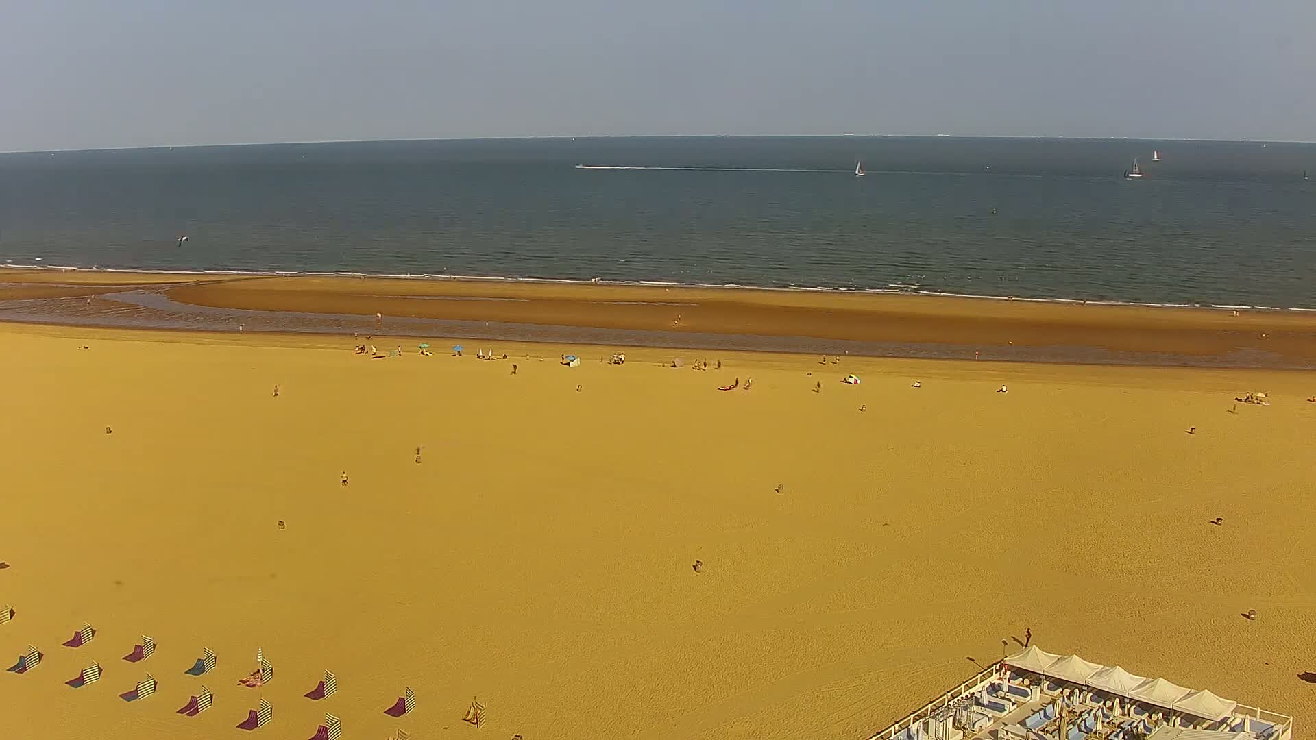 An aerial view shows a wide sandy beach with scattered sunbathers, beach umbrellas, and striped beach chairs, bordered by a calm sea with a few sailboats on a sunny day.