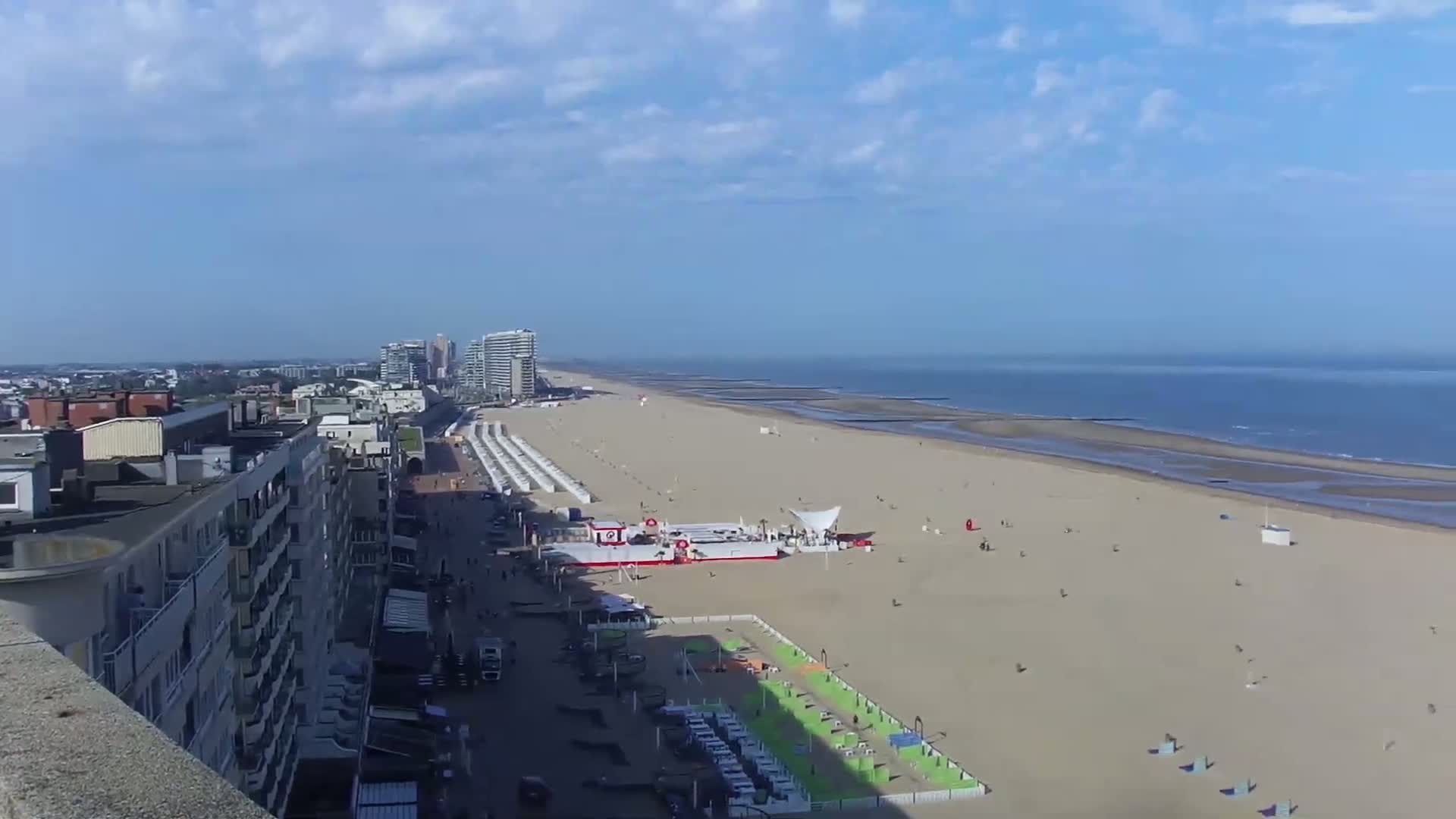 A mostly sunny day shows a long sandy beach with a few people, beach structures, and buildings along the coast, overlooking a calm ocean.