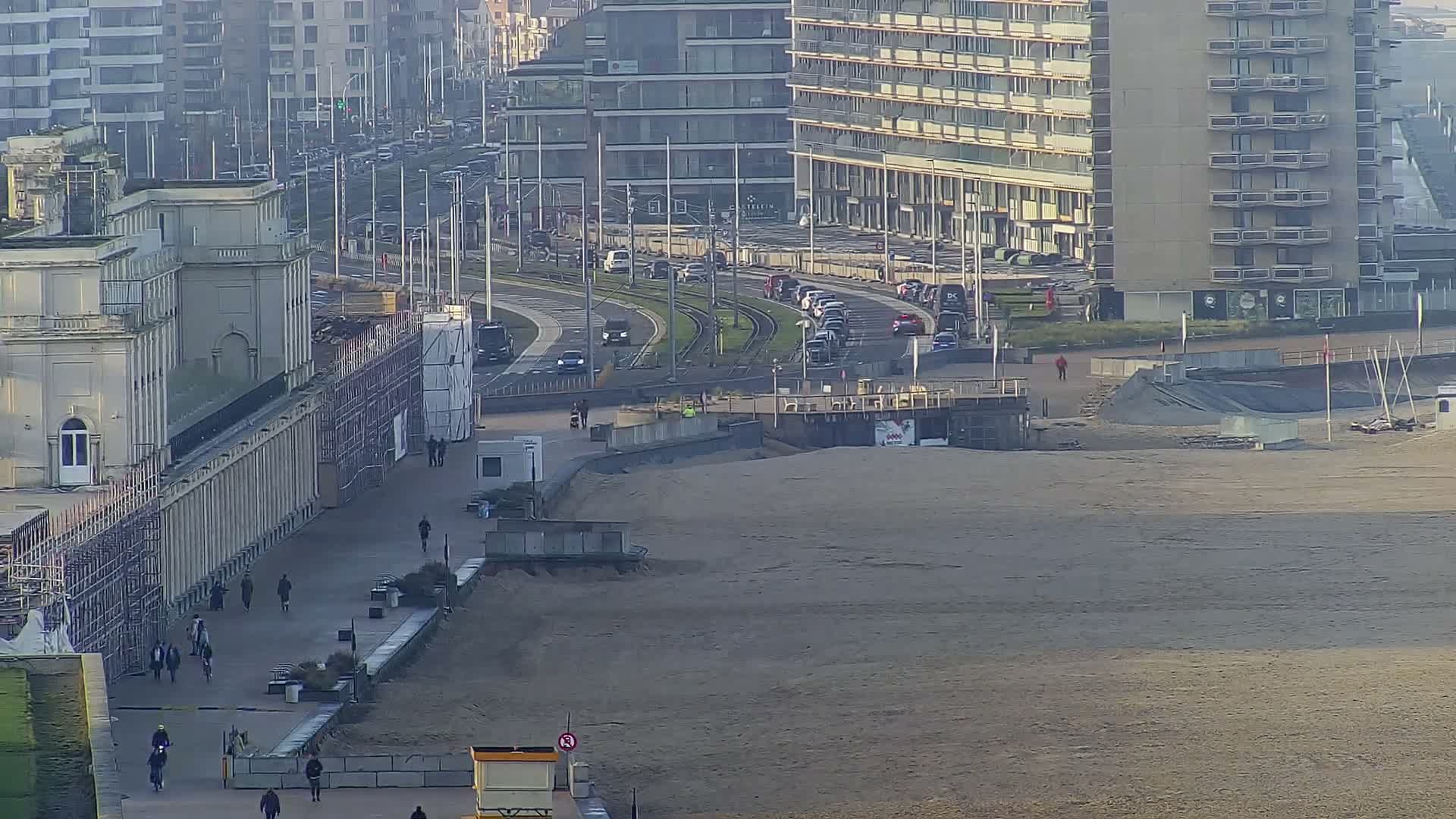 On a hazy day, a wide sandy beach stretches alongside a promenade populated by pedestrians and cyclists, a curved multi-lane road carrying cars and tram tracks, and a dense backdrop of urban buildings.