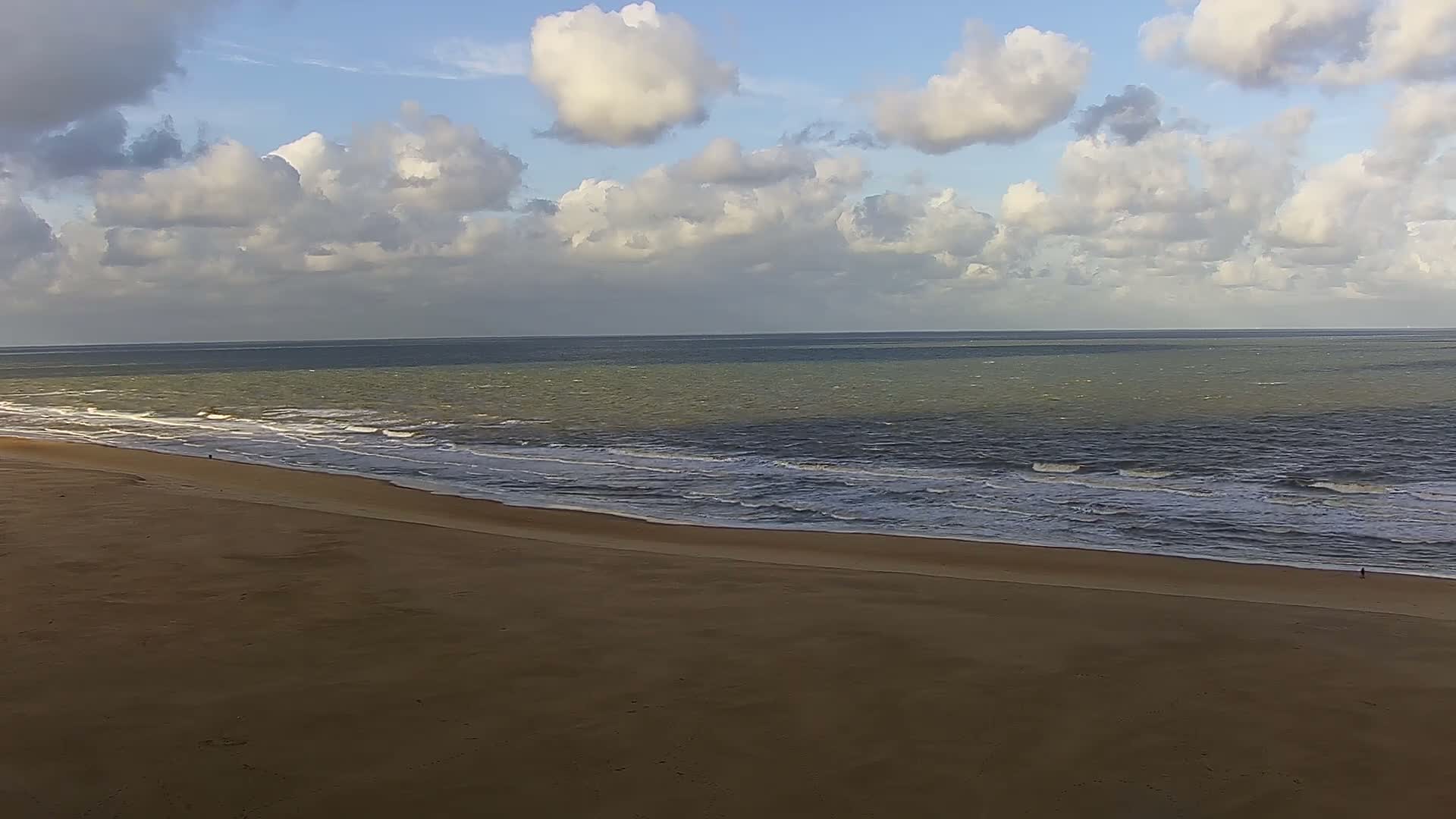 On a hazy day, a wide sandy beach stretches alongside a promenade populated by pedestrians and cyclists, a curved multi-lane road carrying cars and tram tracks, and a dense backdrop of urban buildings.