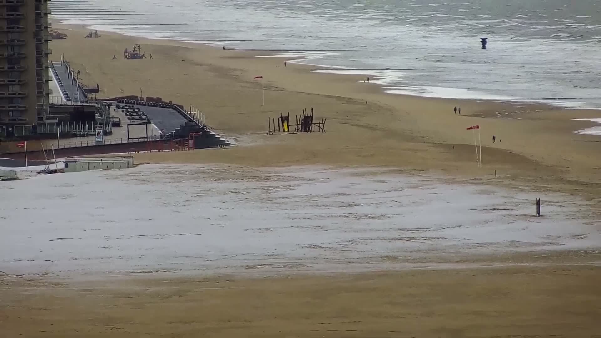 A wide, sandy beach covered in patches of snow and frost is visible on an overcast day, with a tall building, an elevated promenade, and a few people walking near the choppy grey ocean.