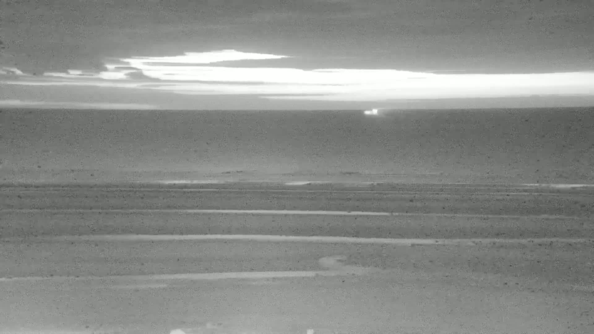 A grayscale image shows a calm body of water under a partly cloudy, bright sky at either dawn or dusk.