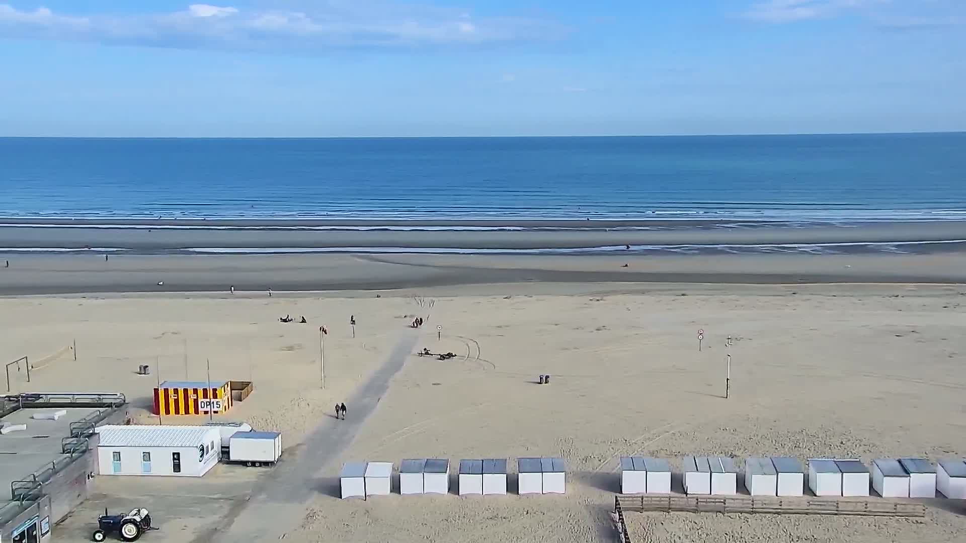 A wide sandy beach with a calm ocean under a mostly clear sky shows small groups of people scattered across the sand near beachside structures and a tractor.