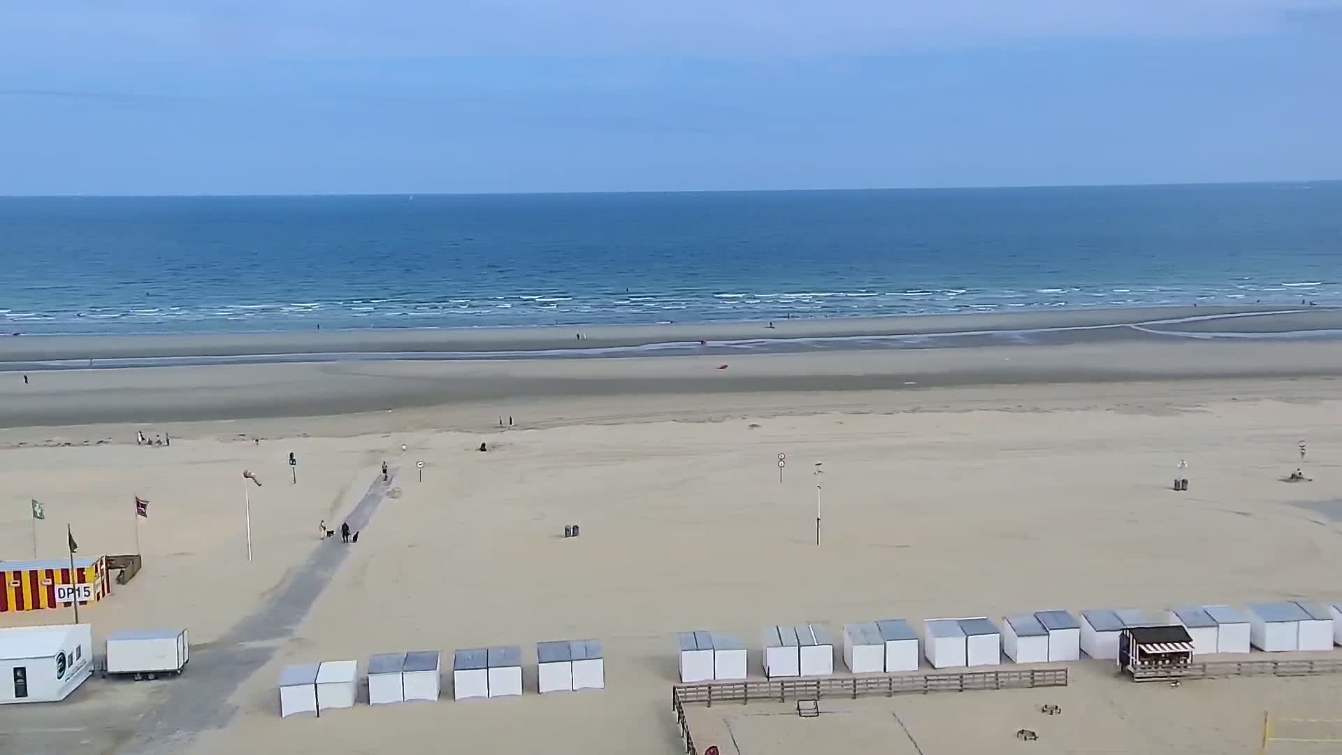 A wide sandy beach with small beach huts and a calm ocean under a mostly clear sky is seen from an elevated perspective.