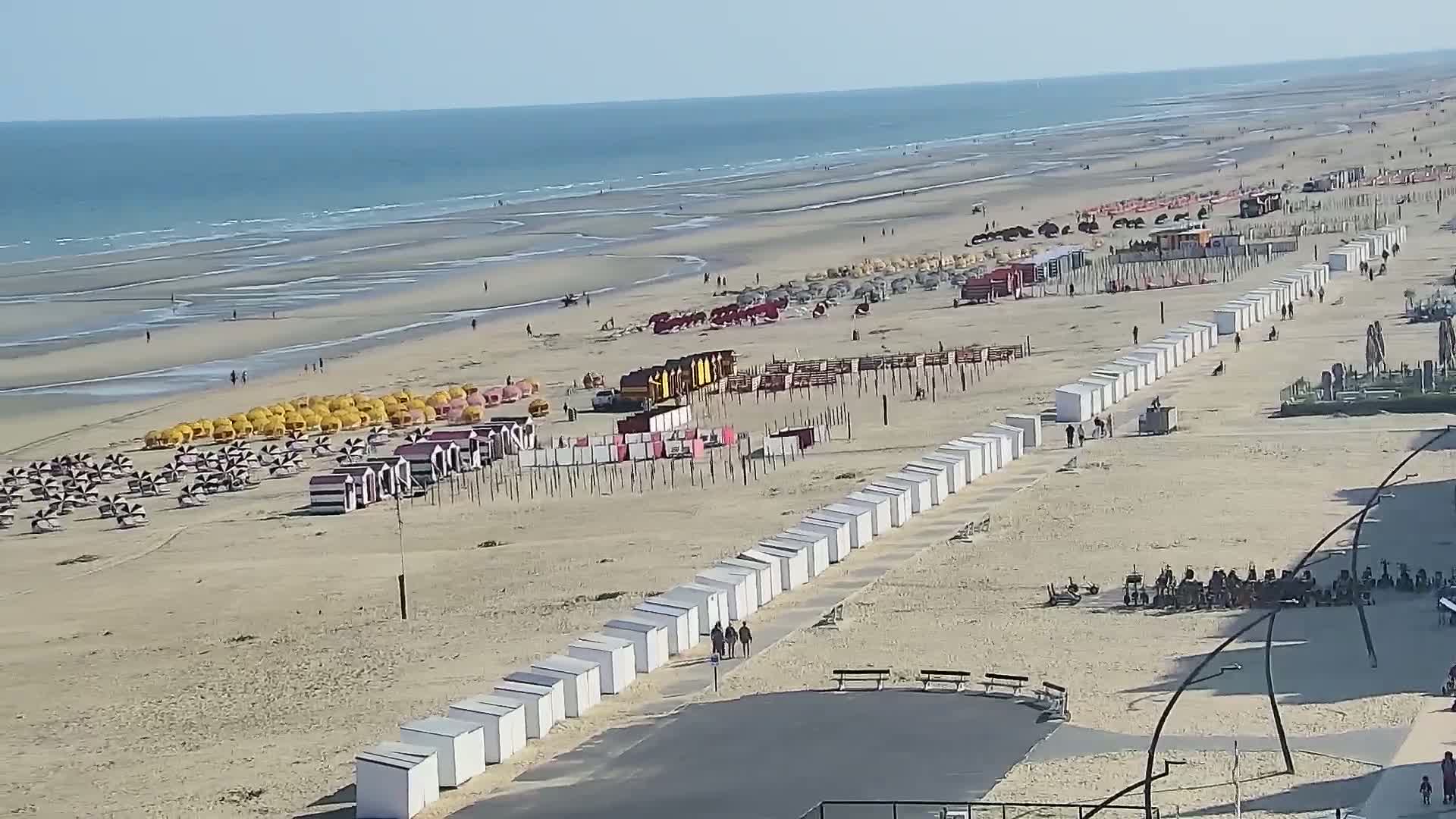 A sandy beach with rows of beach huts and umbrellas stretches towards a calm ocean under a clear, sunny sky.