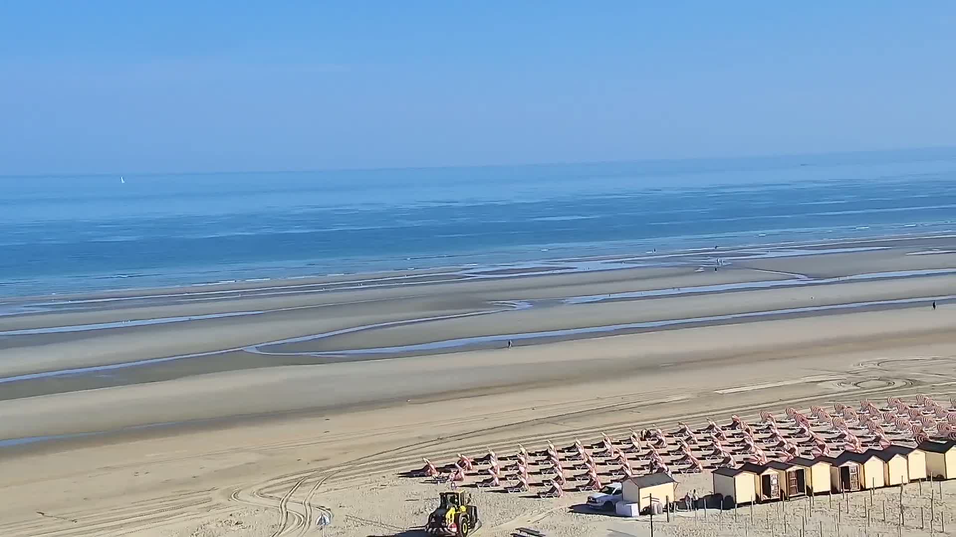 A sandy beach with rows of beach chairs and small huts is shown under a clear blue sky, with a calm ocean visible in the background.