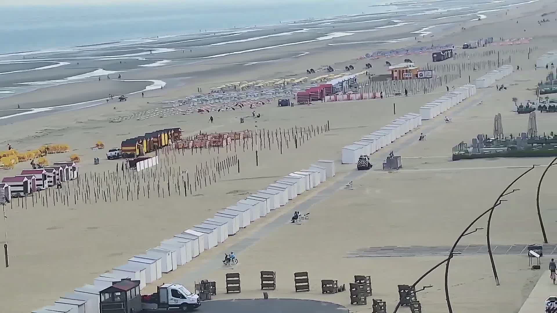 A sandy beach with rows of beach huts and deck chairs, vehicles, and a few people, under an overcast sky and with low tide visible in the ocean.