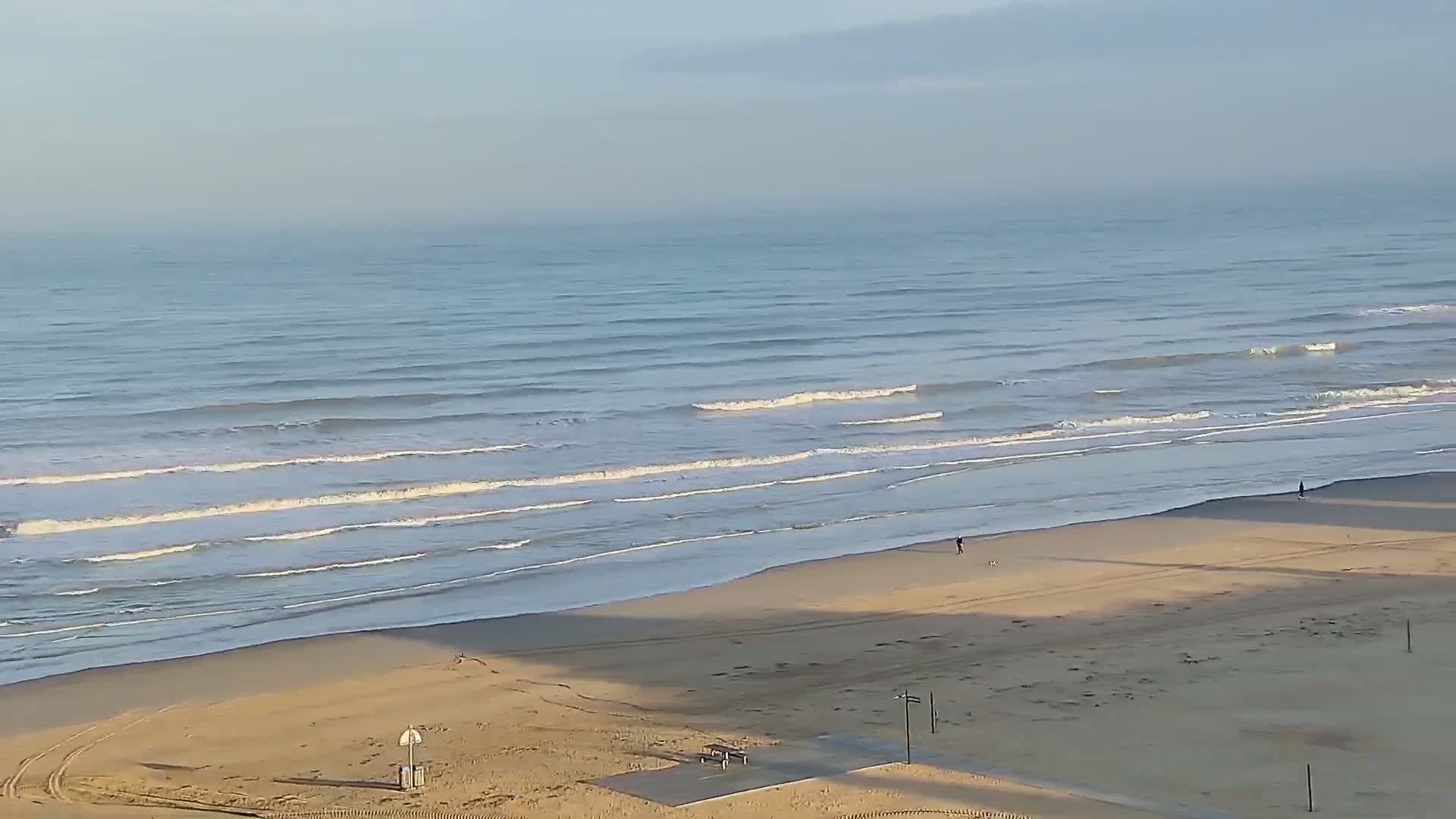 A high-angle view captures a wide sandy beach with gentle waves breaking on the shore, where a few individuals stroll under a bright, partially cloudy sky.