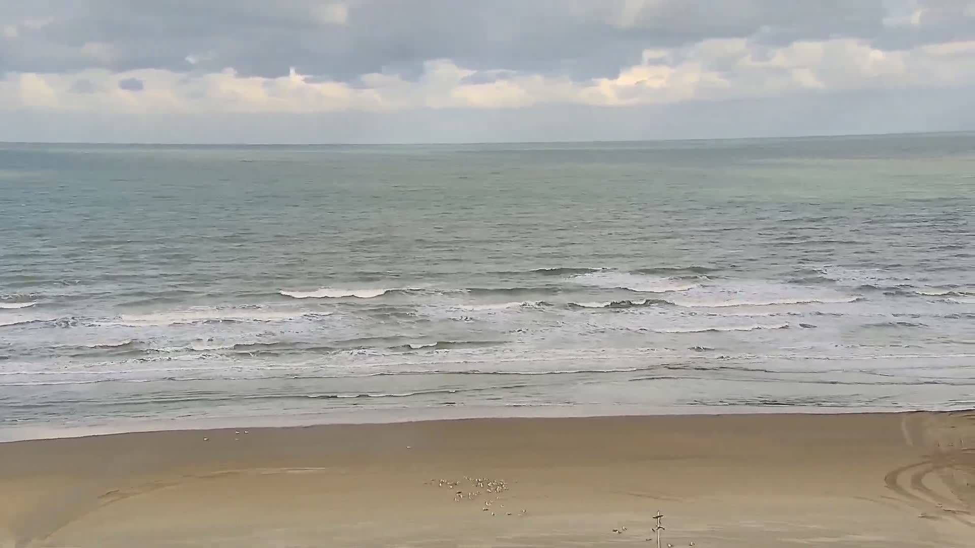 A high-angle view captures a wide sandy beach with gentle waves breaking on the shore, where a few individuals stroll under a bright, partially cloudy sky.