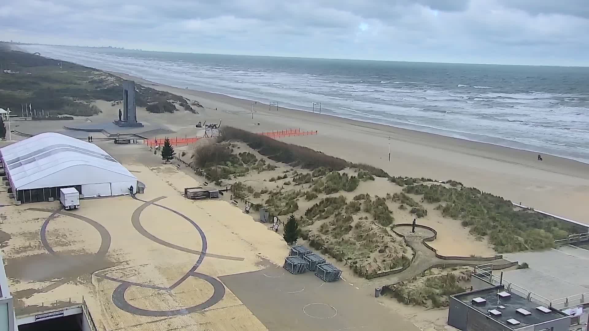 An overcast day reveals a wide sandy beach with active waves, backed by dunes, a large white tent, and a monument, with a few people visible.