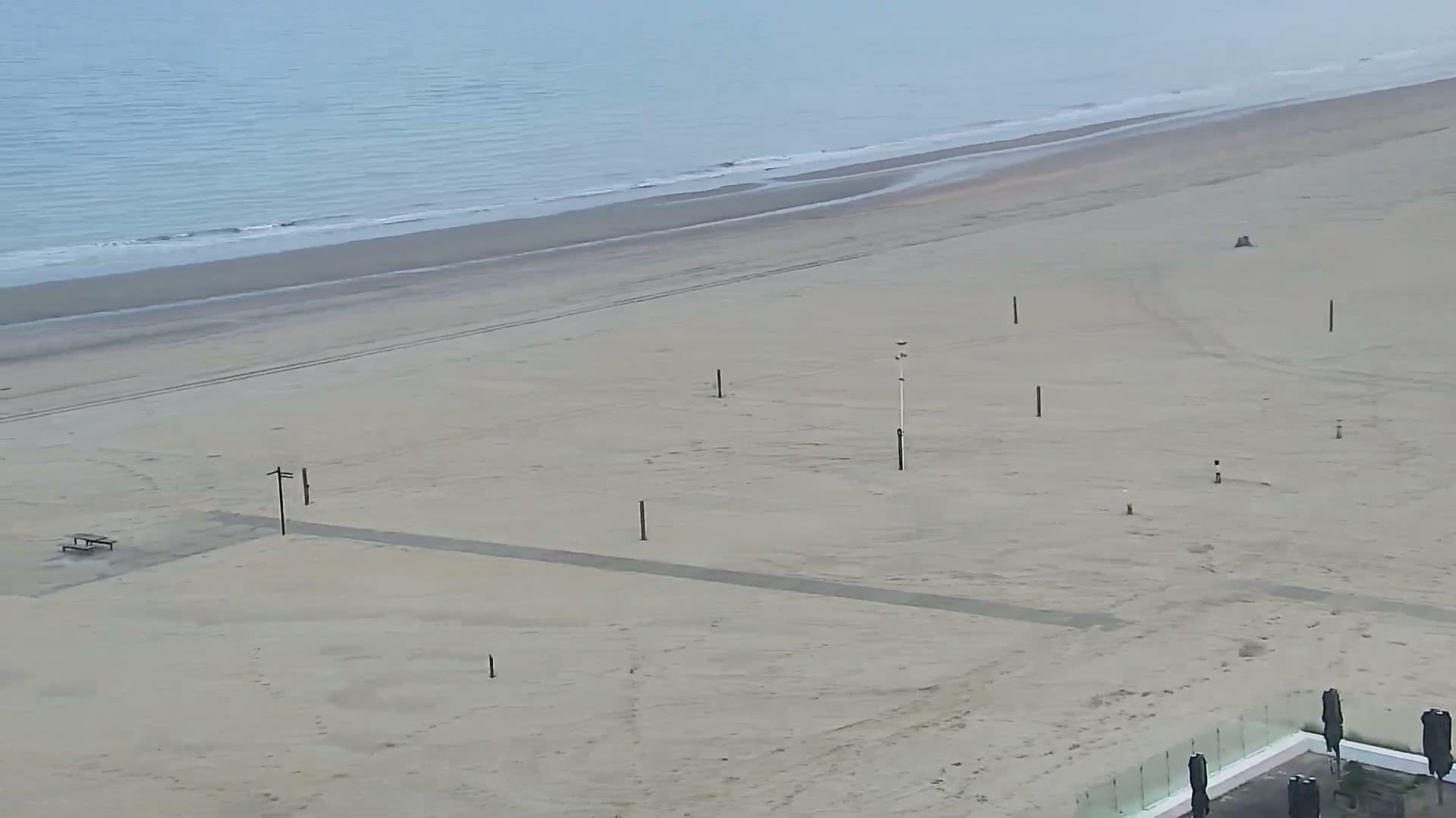 An expansive, mostly empty sandy beach with several scattered dark poles and a small bench leads to calm, light blue ocean water under a soft, overcast sky, with a modern building's terrace partially visible in the bottom right.