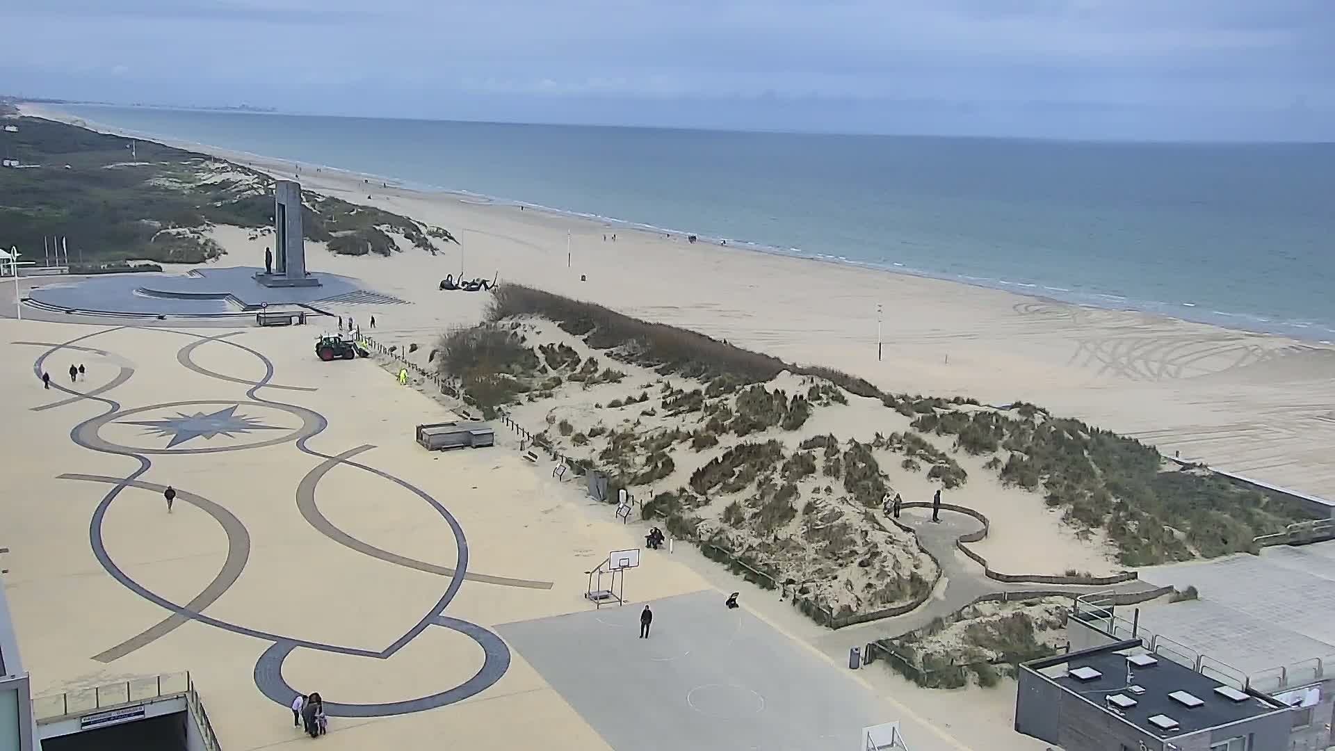An expansive, mostly empty sandy beach with several scattered dark poles and a small bench leads to calm, light blue ocean water under a soft, overcast sky, with a modern building's terrace partially visible in the bottom right.