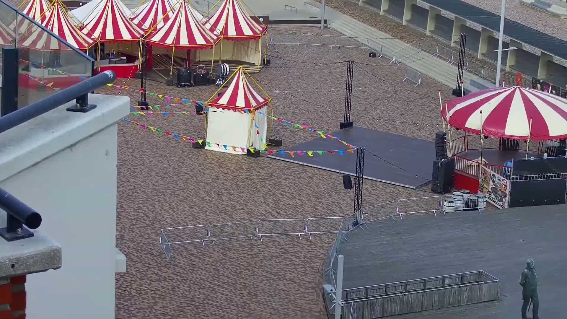 Several red and white striped circus tents and stages are set up in an outdoor paved area, decorated with colorful flags.