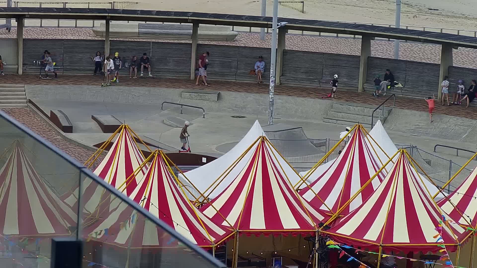 A skate park with several people on scooters and skateboards is adjacent to a cluster of red and white striped circus tents under an overcast sky.