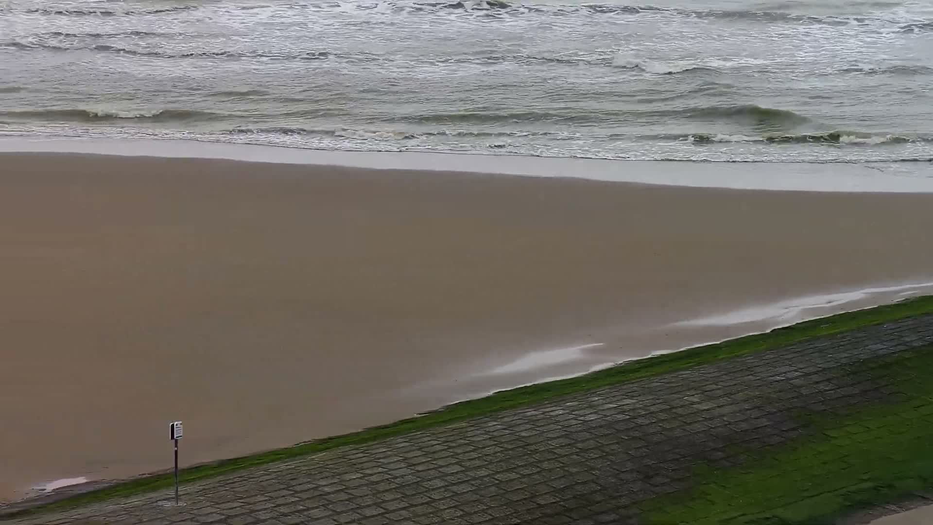 An overcast day reveals a wide, empty sandy beach bordered by a choppy grey ocean and a moss-covered, tiled seawall with a small, indistinct sign.