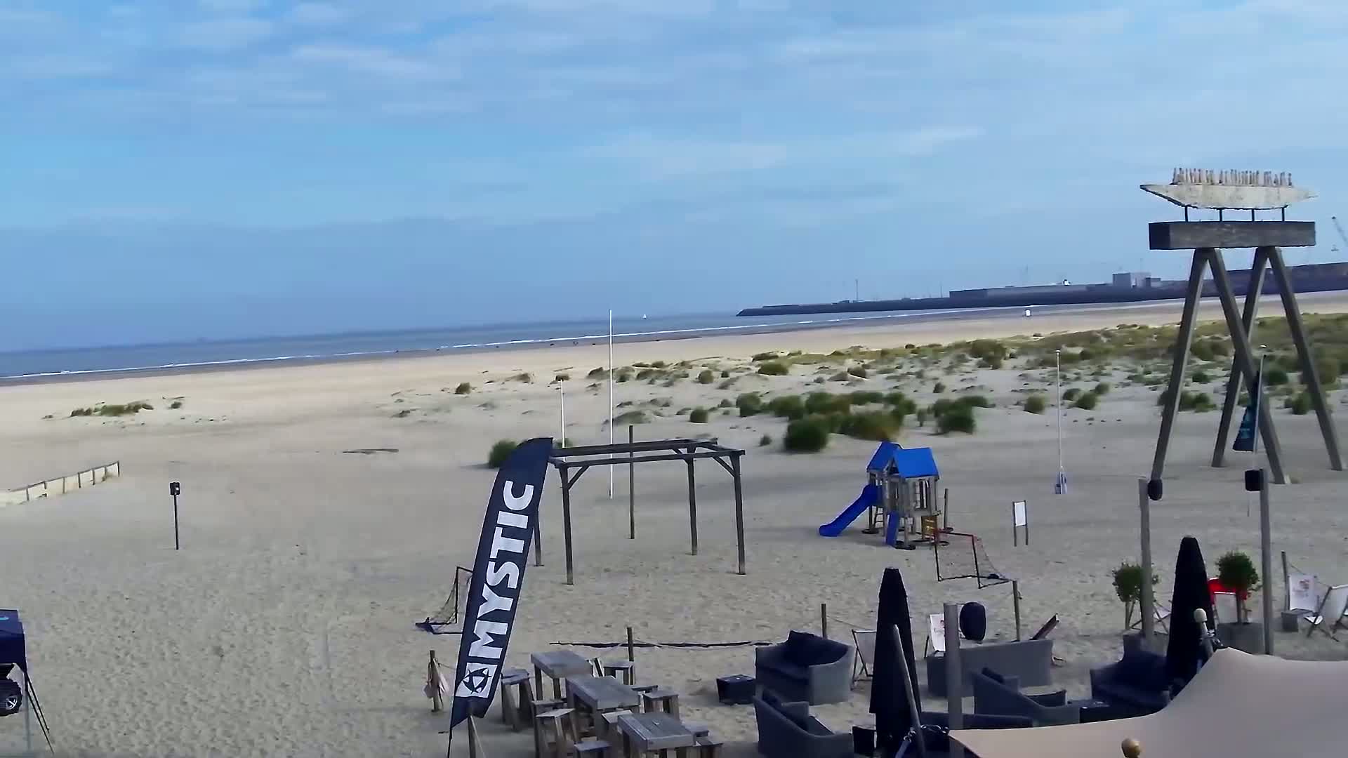 A sandy beach with a playground, outdoor furniture, and a calm ocean under a mostly clear sky is visible.