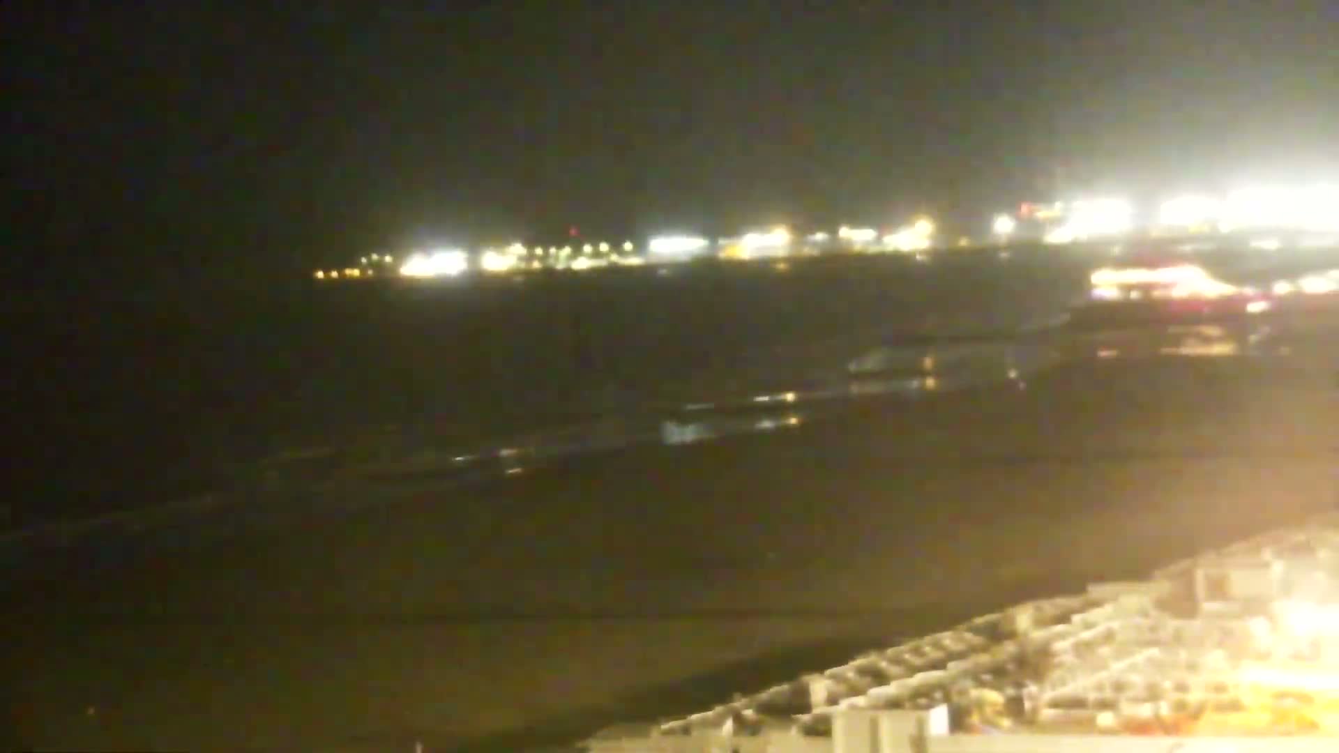 A dark beach at night is shown with a distant city skyline illuminated by lights, and gentle waves lapping the shore.