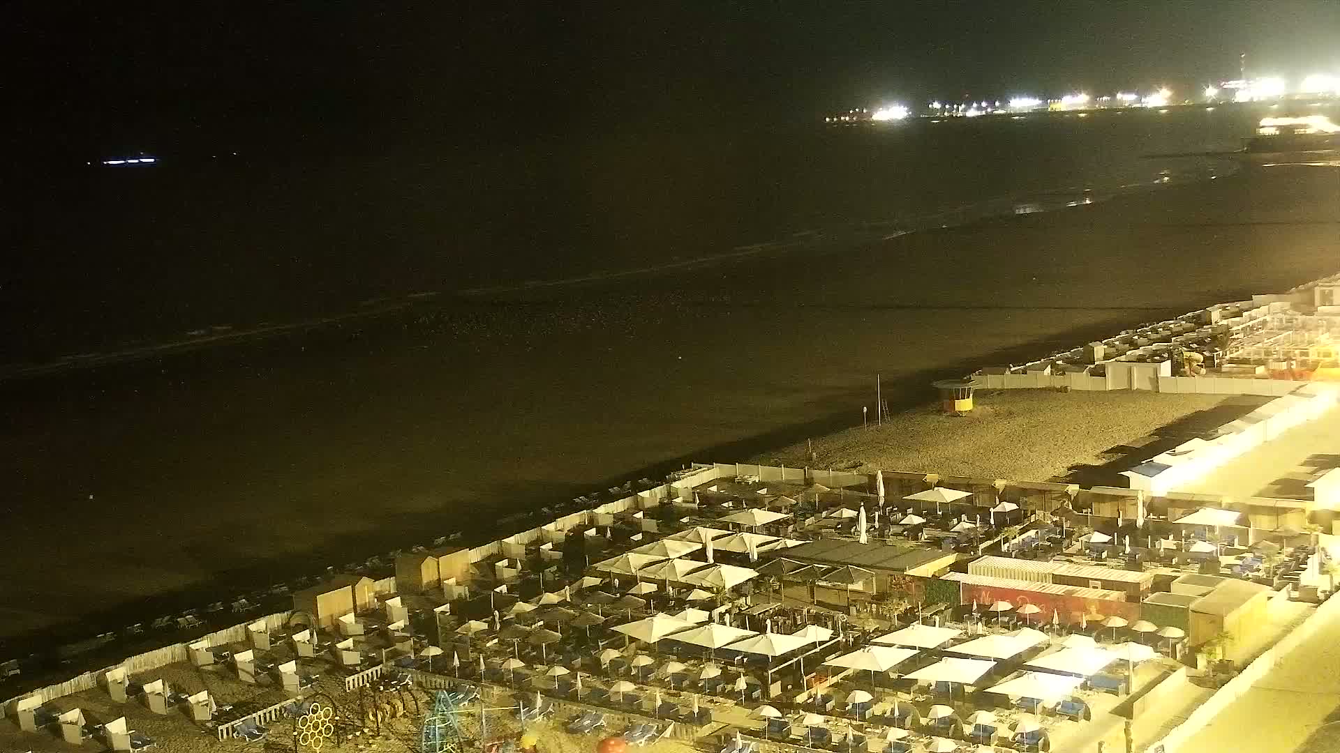 A dark beach at night is shown with a distant city skyline illuminated by lights, and gentle waves lapping the shore.