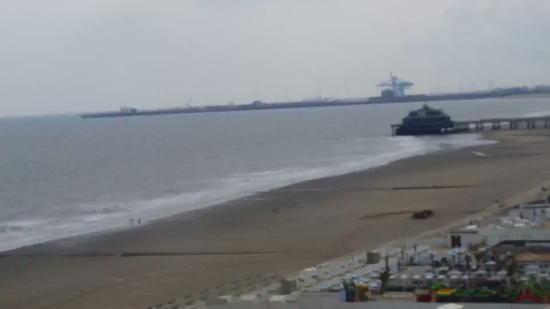 A beach with a pier extending into a calm, grey sea under an overcast sky, featuring some beach structures in the foreground.