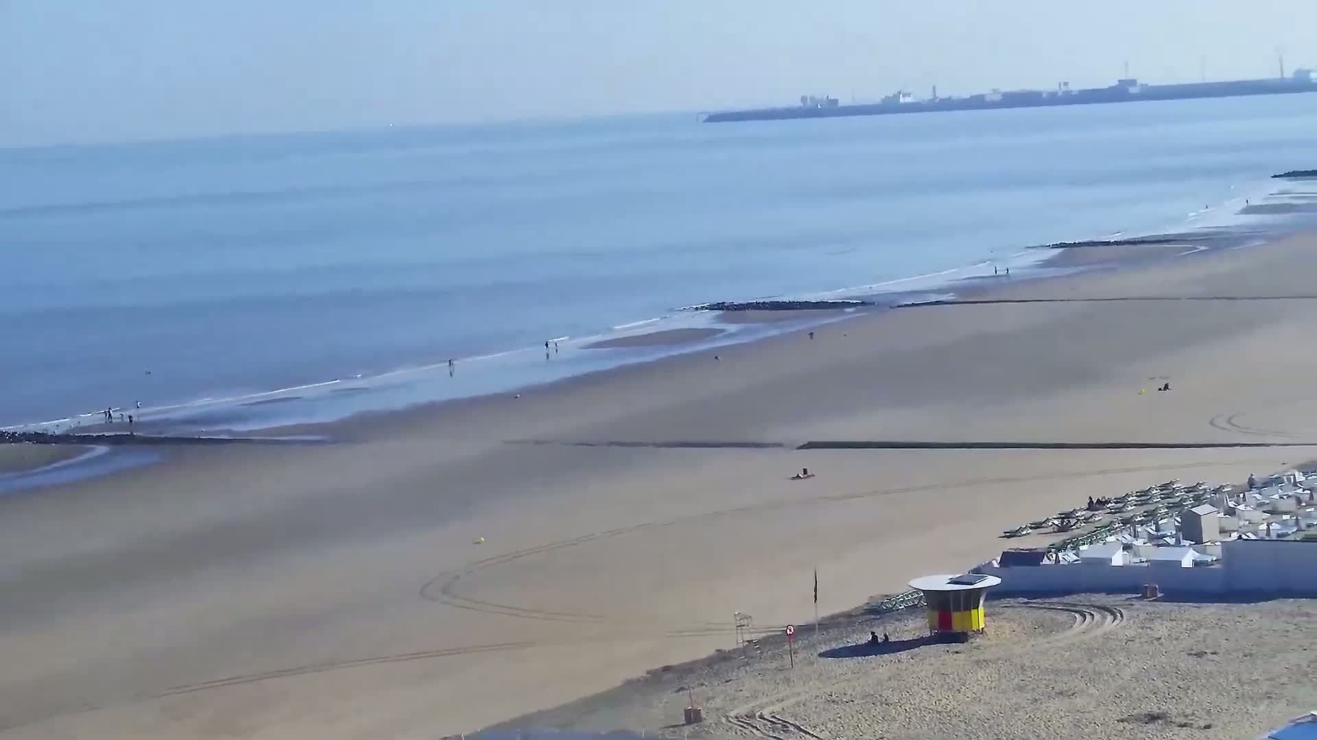 A long sandy beach stretches toward a calm ocean under a clear sky, with a few people scattered along the shore and beach structures near the lower right corner.