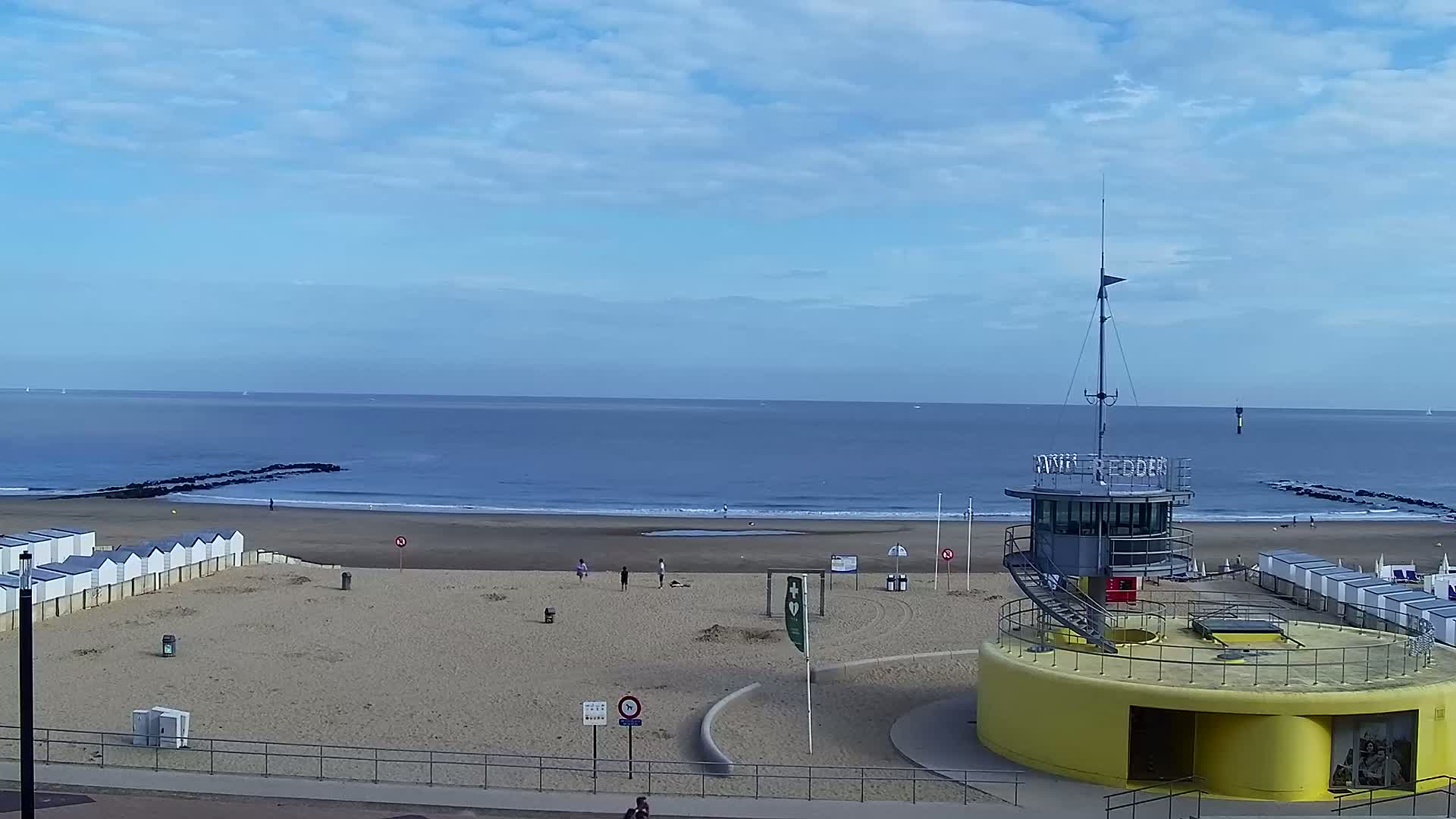 A sandy beach with rows of small white beach huts, a yellow lifeguard tower, and a calm sea under a partly cloudy sky.
