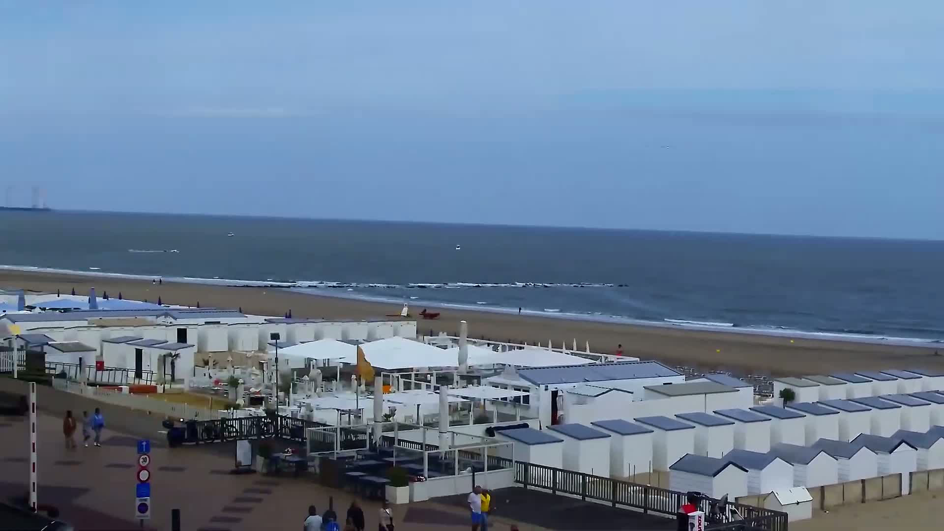 A sandy beach with calm ocean waves, rows of white beach huts, and outdoor seating areas is seen from a high vantage point on a partly cloudy day.