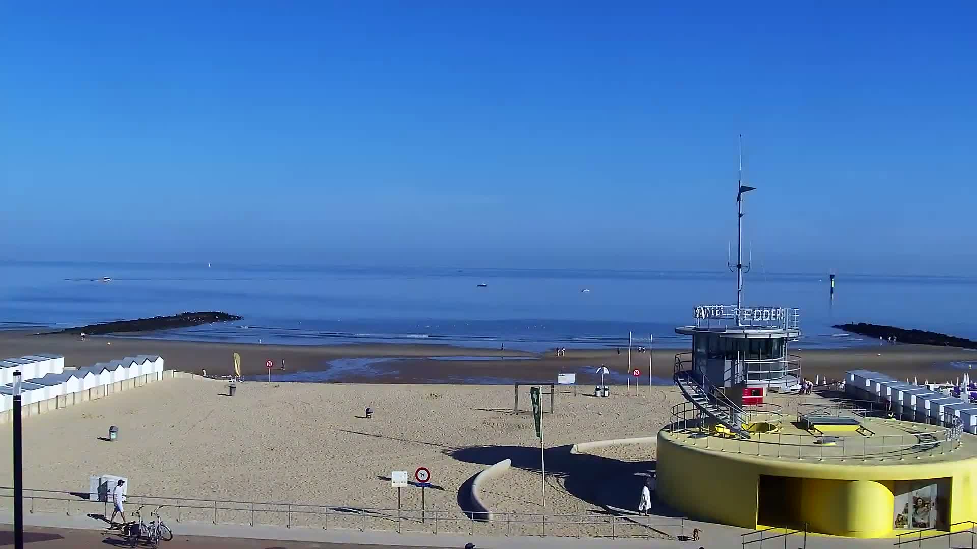 A sandy beach with white beach huts, a lifeguard tower, and a calm, clear blue sea under a bright blue sky.