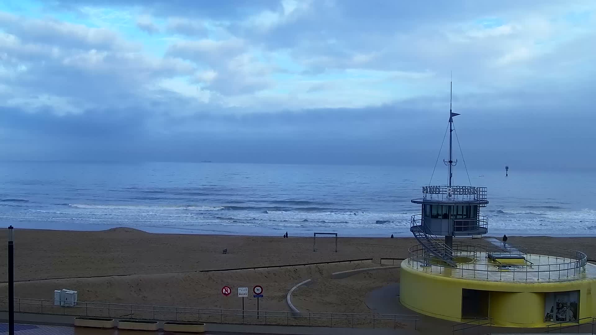 An overcast sky hangs above a wide, sandy beach where moderate waves break into the blue ocean, featuring a prominent yellow and grey circular building with a lookout tower on the right, a distant ship on the horizon, and a few sparse figures on the sand.