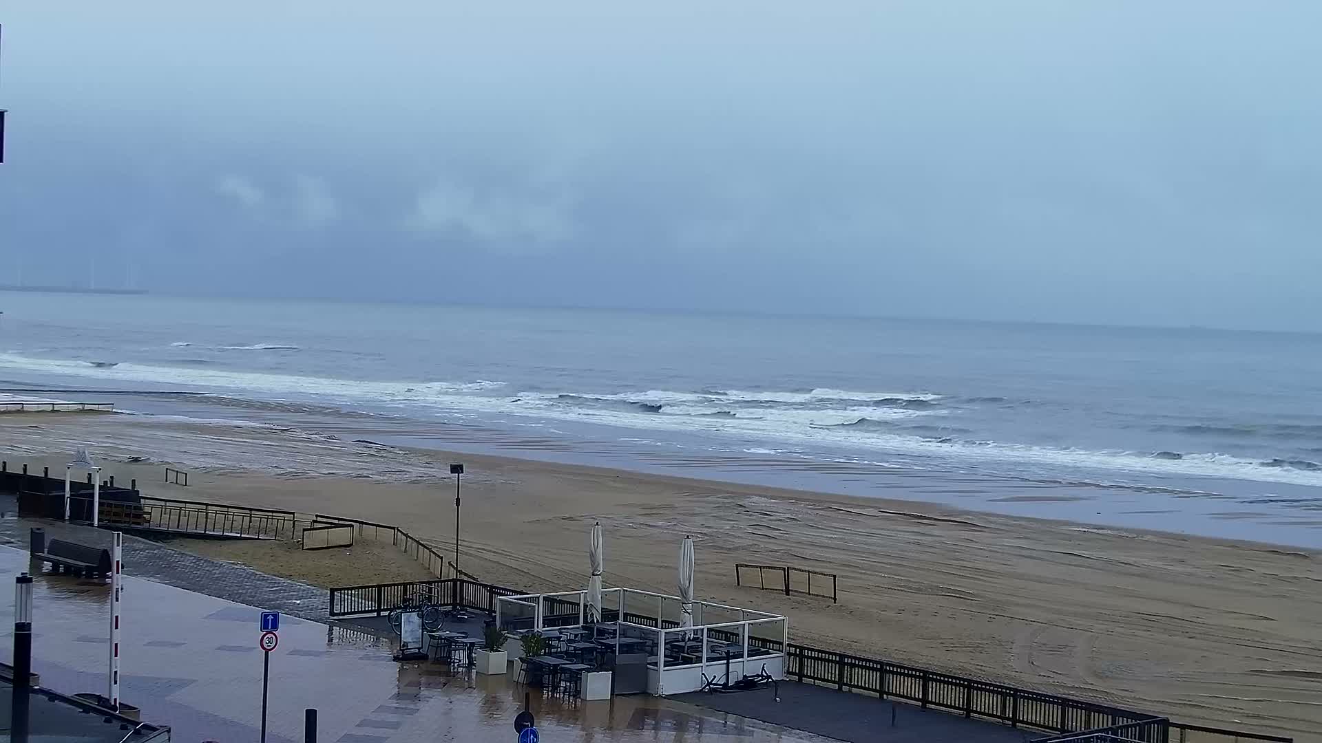 A gloomy, overcast, and rainy day is depicted at a deserted beach, with grey waves breaking on the wide sandy shore, and a wet paved area with an empty outdoor cafe terrace in the foreground.