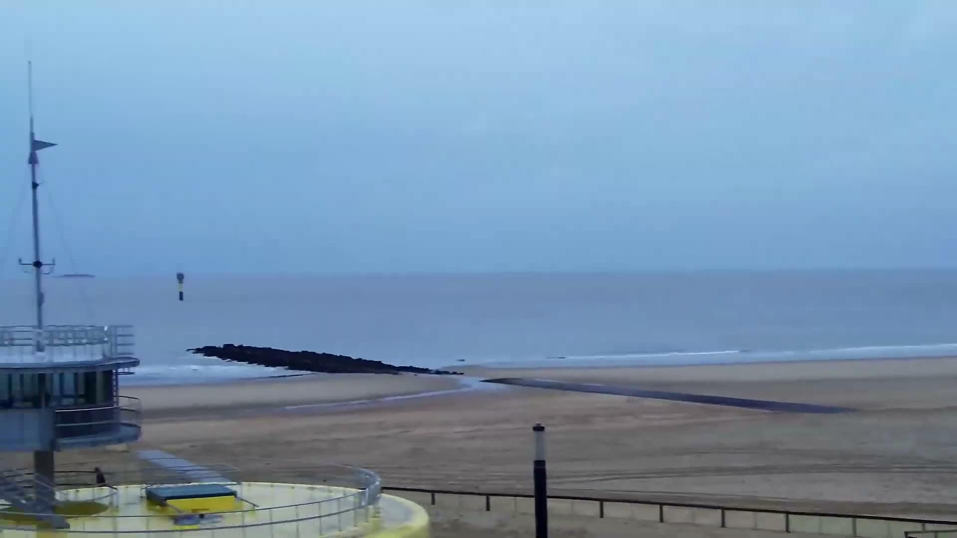 Under an overcast sky, a tranquil sandy beach with a rock groyne meets a calm ocean, viewed from the upper level of a modern, circular building with a mast on the left.