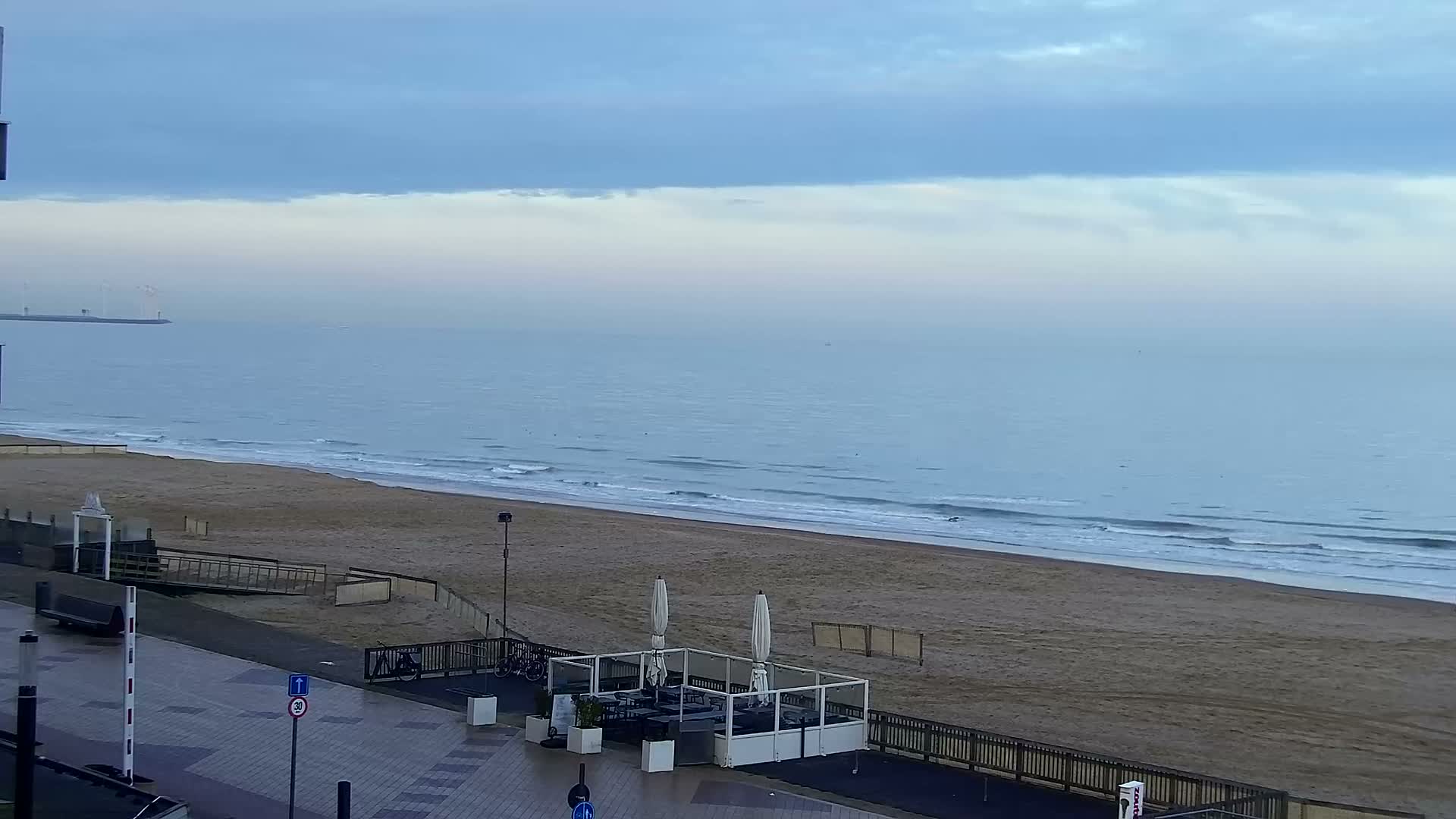Under an overcast blue-gray sky, a peaceful wide sandy beach with gentle waves meets a calm ocean, overlooked by a paved promenade featuring a fenced-off outdoor dining area and a distant pier with structures on the left.