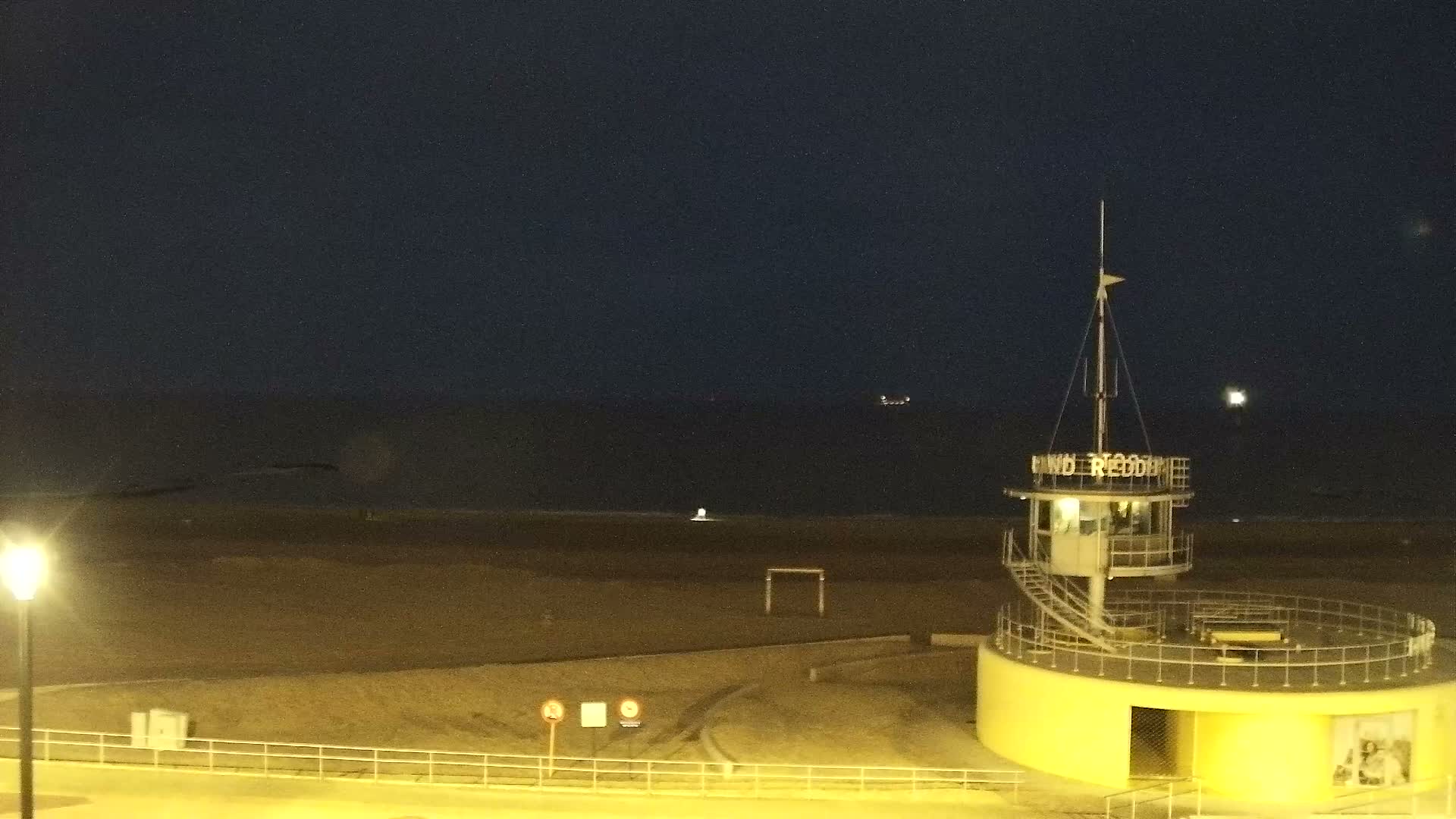 Under an overcast blue-gray sky, a peaceful wide sandy beach with gentle waves meets a calm ocean, overlooked by a paved promenade featuring a fenced-off outdoor dining area and a distant pier with structures on the left.