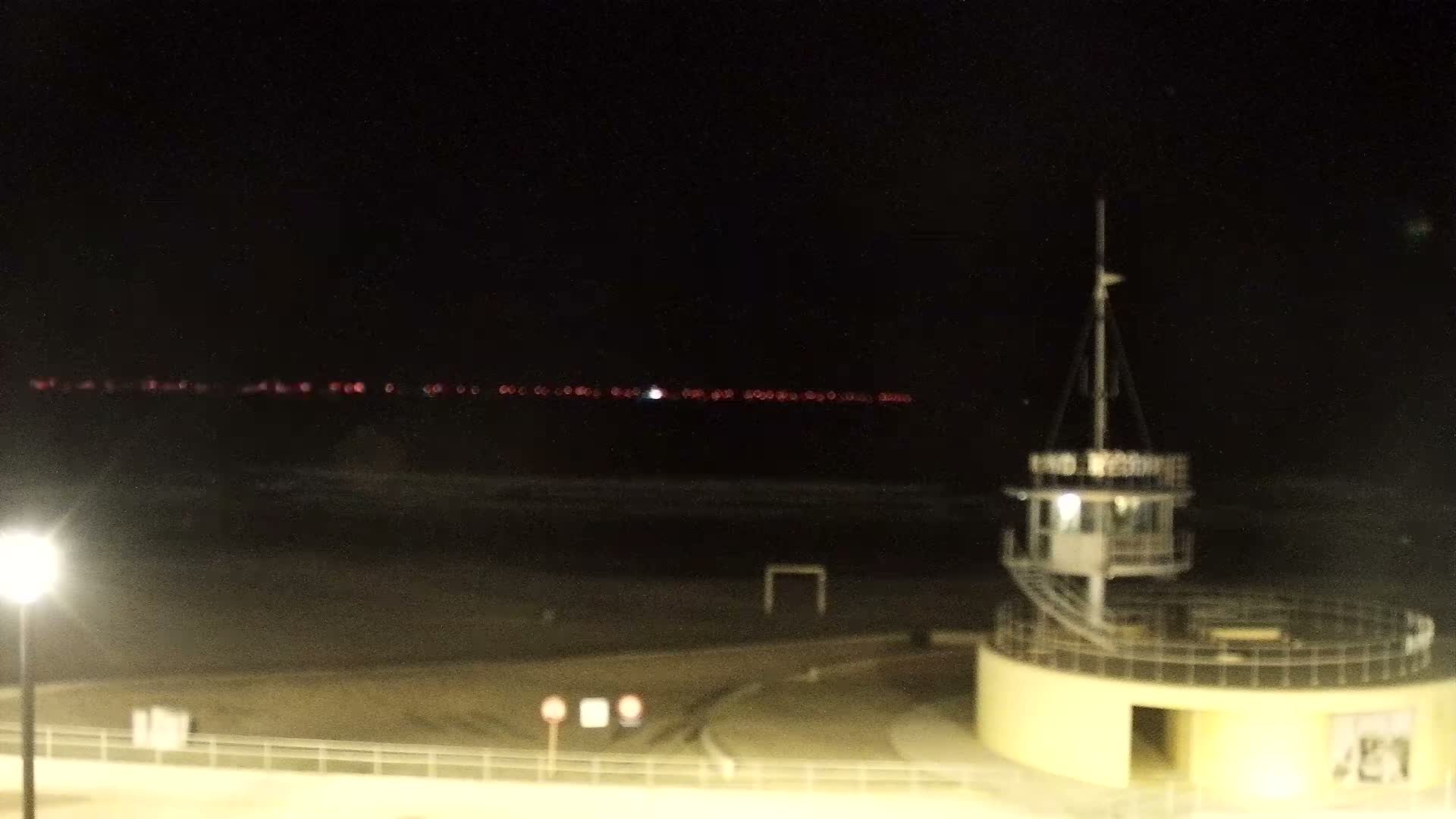 Under an overcast blue-gray sky, a peaceful wide sandy beach with gentle waves meets a calm ocean, overlooked by a paved promenade featuring a fenced-off outdoor dining area and a distant pier with structures on the left.