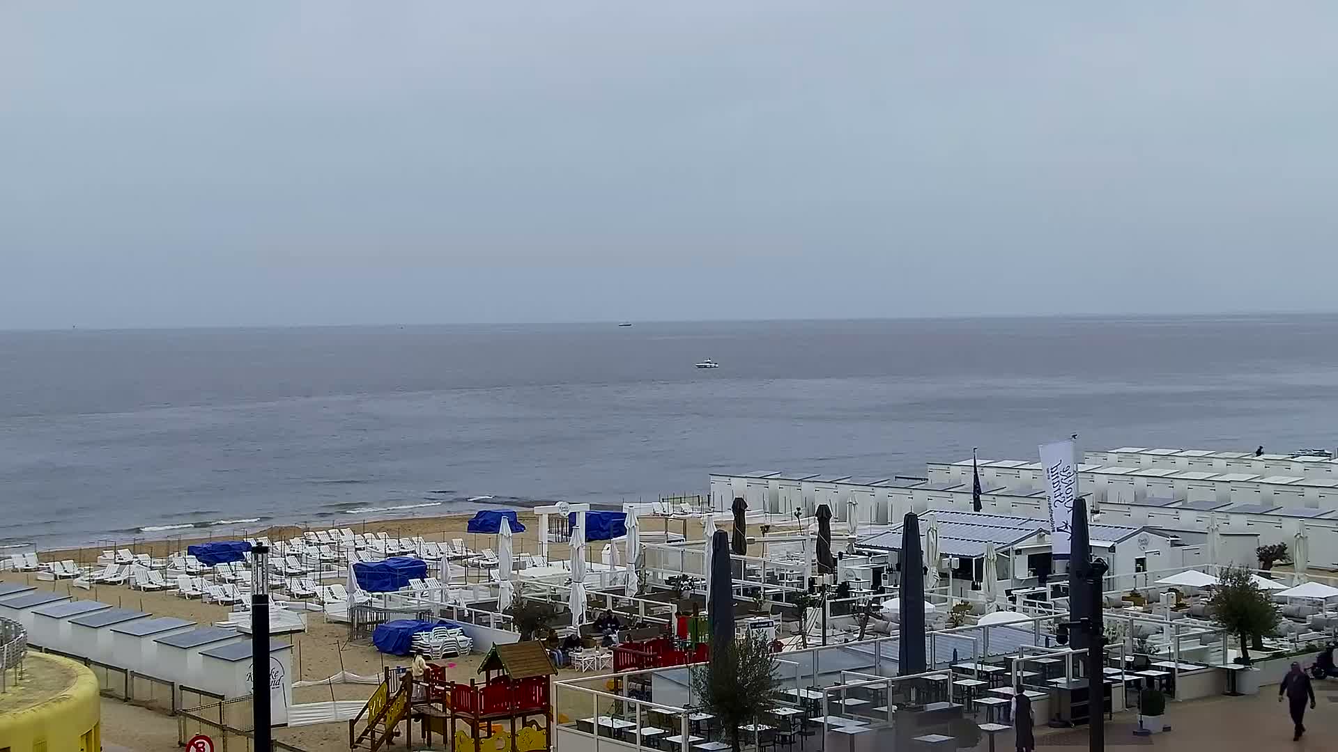 Under an overcast blue-gray sky, a peaceful wide sandy beach with gentle waves meets a calm ocean, overlooked by a paved promenade featuring a fenced-off outdoor dining area and a distant pier with structures on the left.