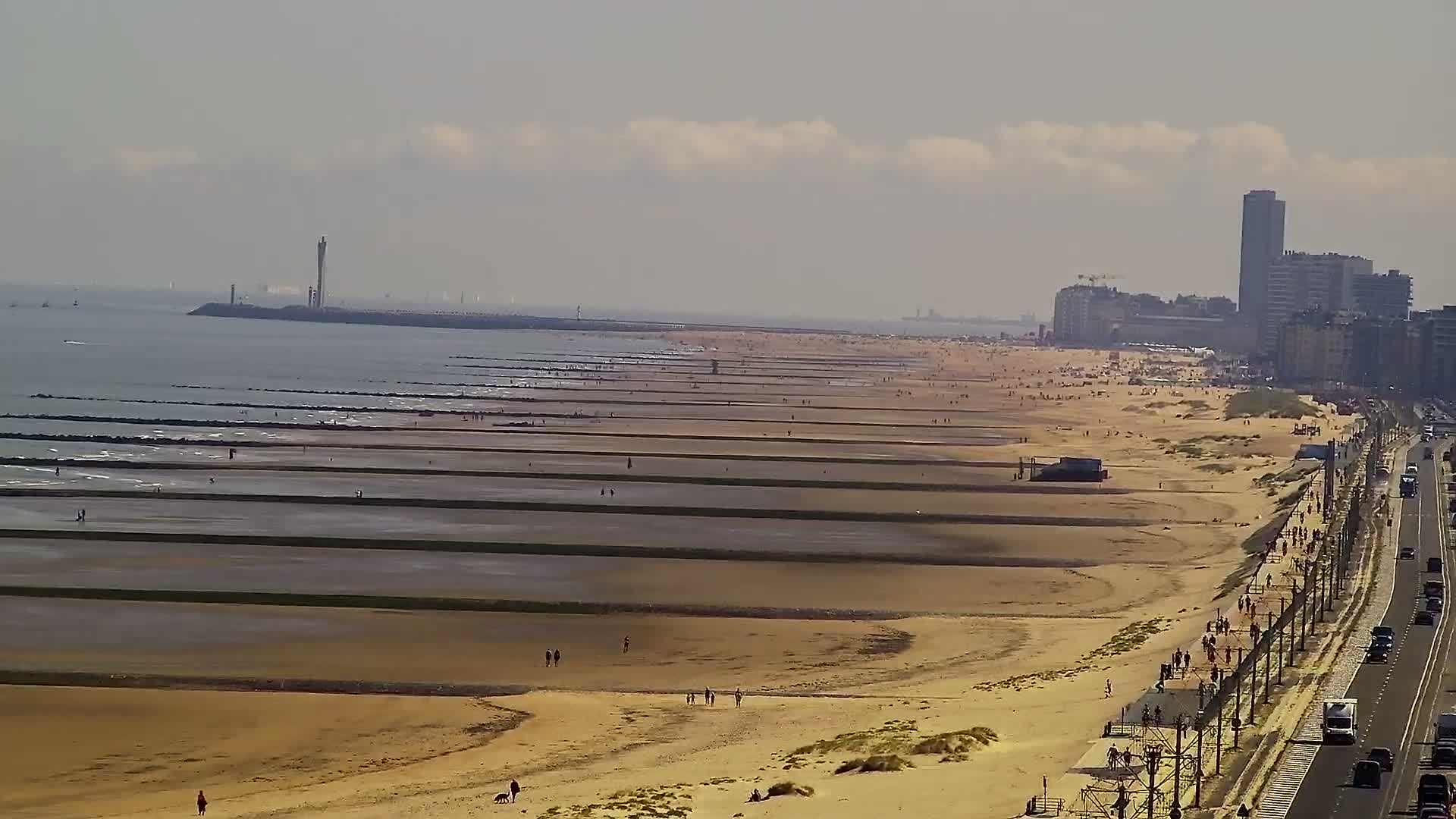 A long sandy beach with many people and parallel structures extending into the water under a partly cloudy sky.