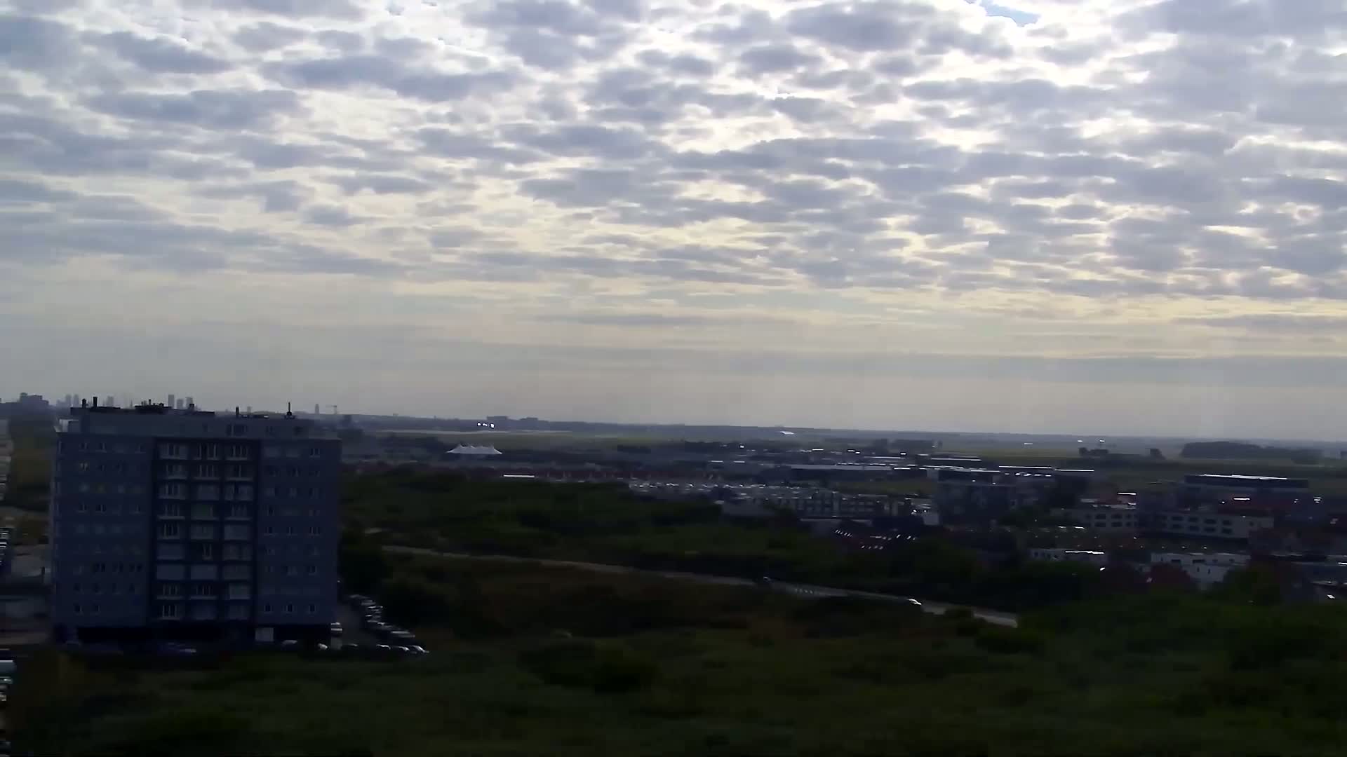 A high-angle, long shot shows a partly cloudy sky over a cityscape, with a multi-story building in the foreground and an airport runway visible in the distance.