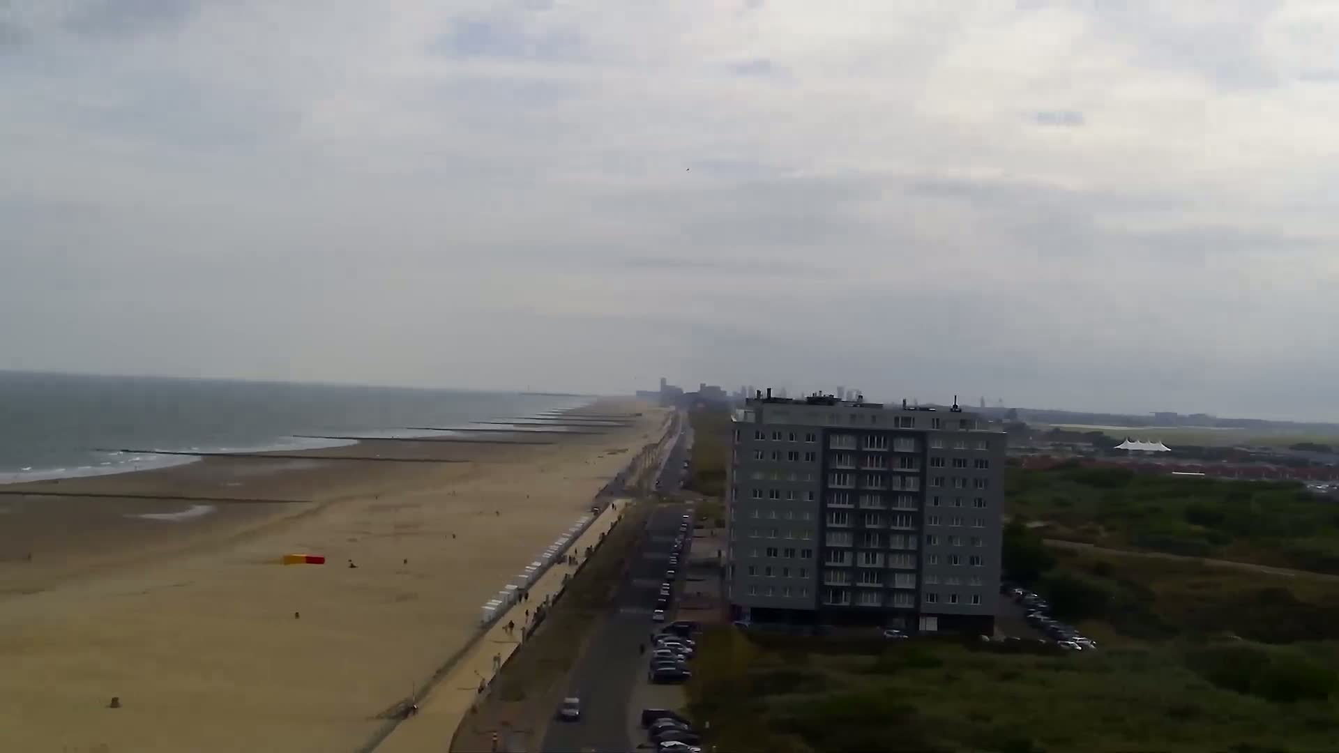 An overcast day reveals a beach with a boardwalk, some people, and breakwaters, extending to a high-rise building and parking lot next to a dune landscape.
