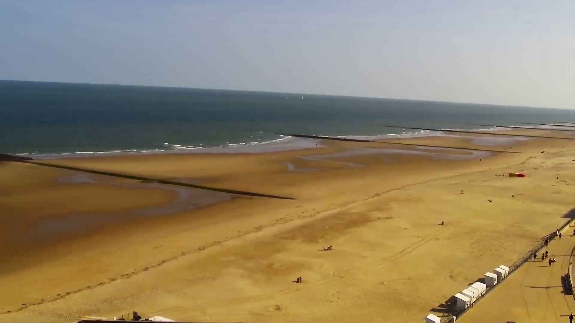 An aerial view shows a wide, sandy beach with several people scattered across it,  under a clear, sunny sky, and a calm sea with small waves breaking gently on the shore, featuring a series of low, dark breakwaters extending into the water.