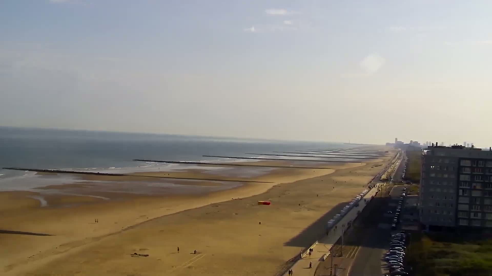A long sandy beach with several breakwaters extends to a calm sea under a hazy sky, next to a paved road lined with cars and a tall apartment building.