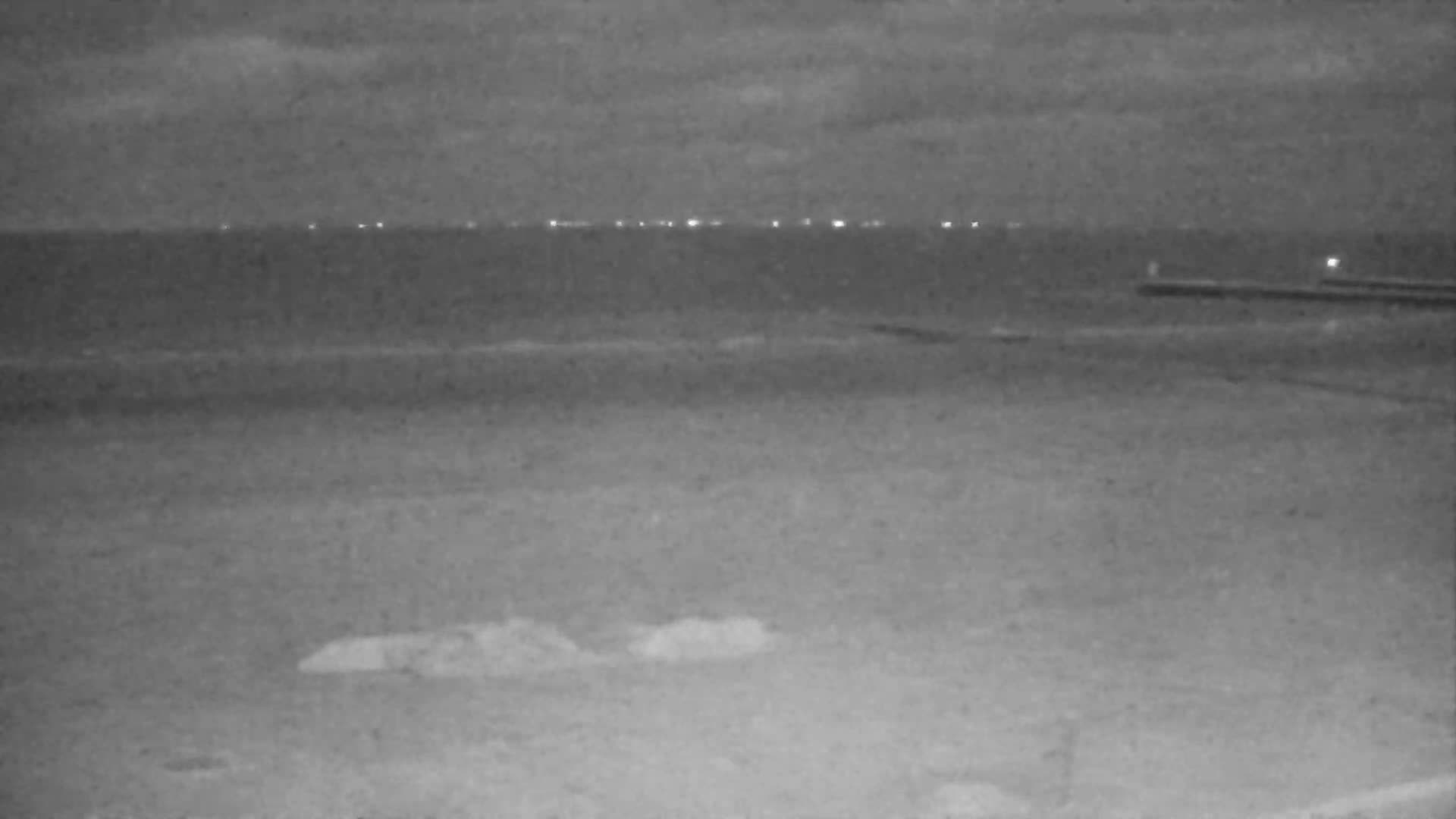 An expansive view captures a calm, gray sea meeting a wide, sandy-muddy beach scattered with dark shapes along the water's edge, all under a heavily overcast and hazy sky.